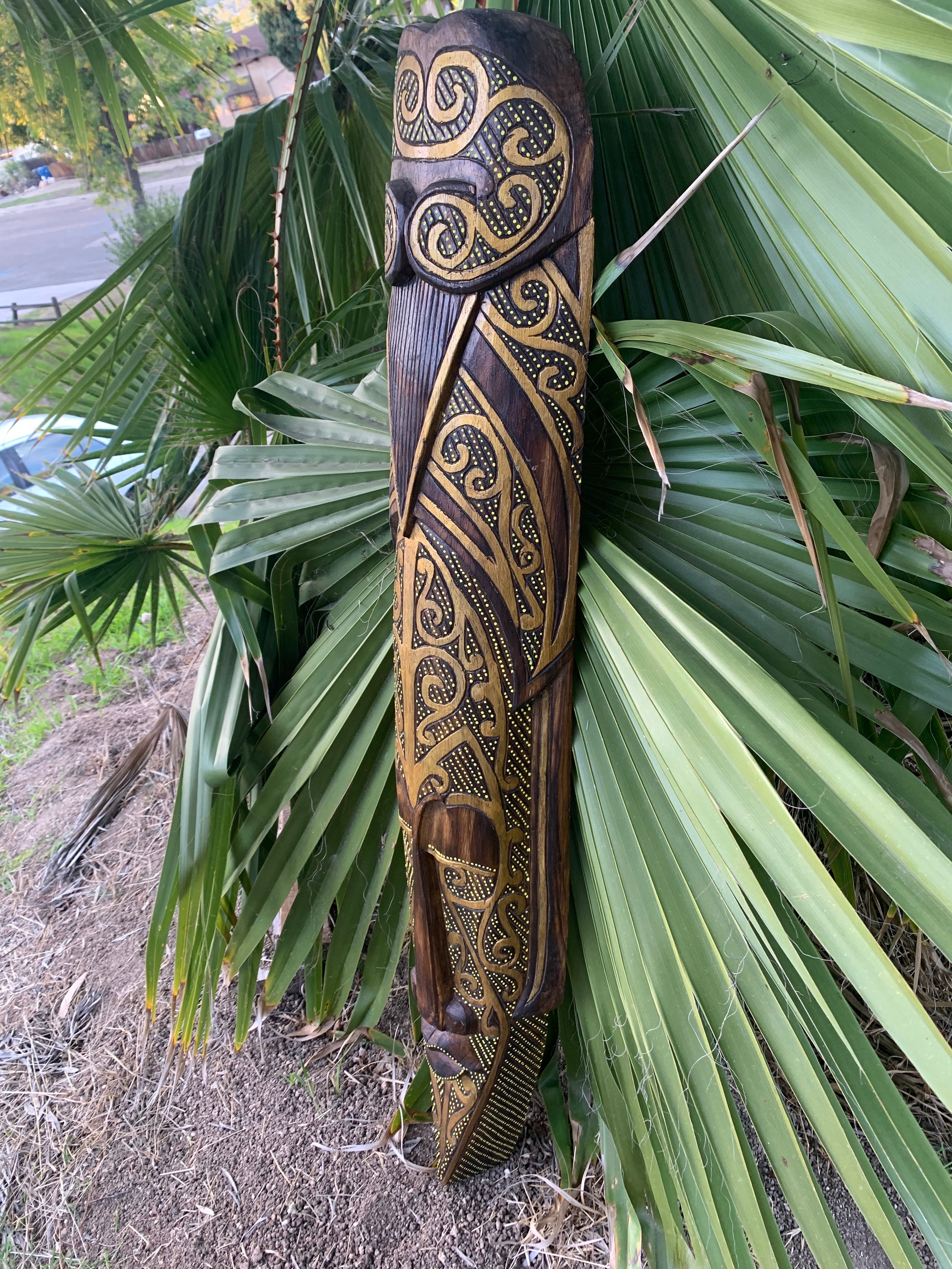 Ornate wooden tiki sculpture with gold scroll patterns leaning against palm leaves