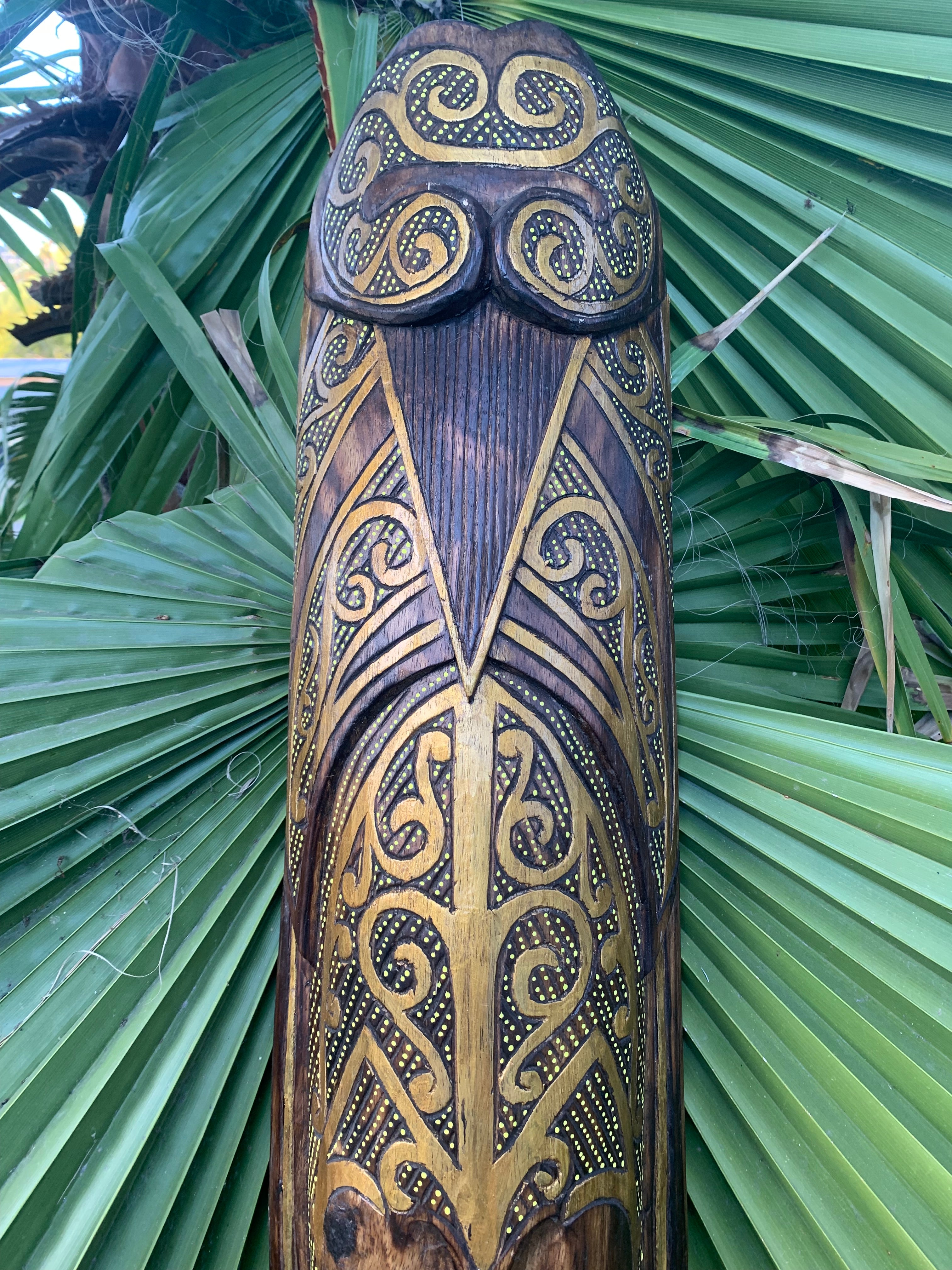 Ornate wooden totem pole with gold scrollwork and dot detailing against green palm leaves.