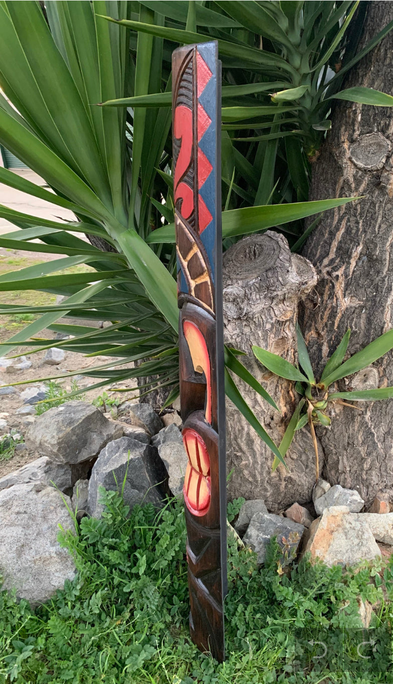 Tall carved wooden totem pole with red geometric patterns outdoors beside greenery