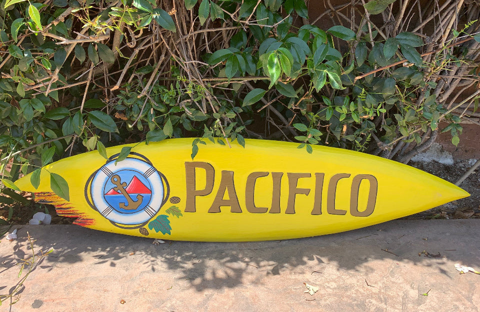 Bright yellow surfboard sign reading PACIFICO with an anchor logo, resting on a sidewalk near green bushes.