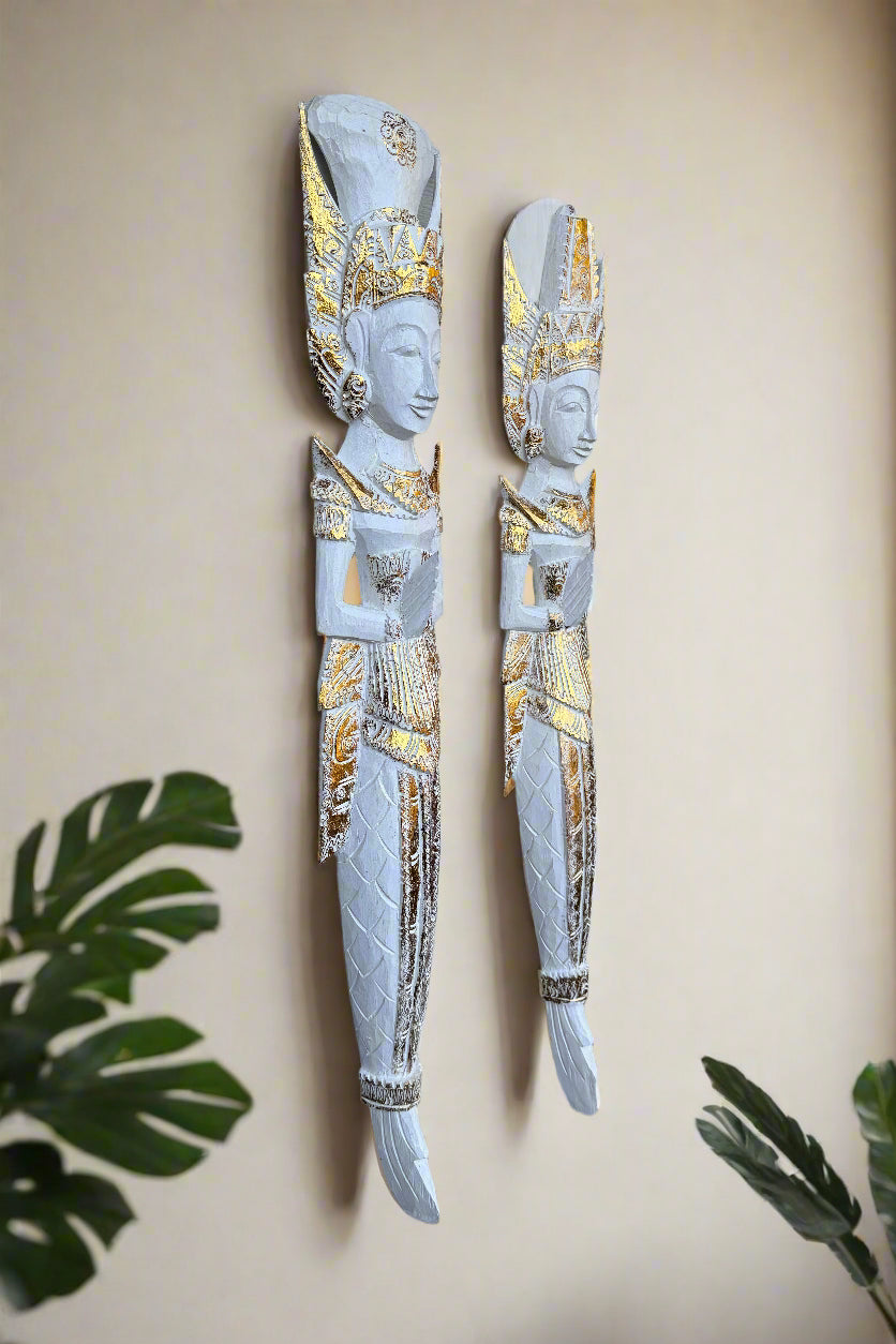 Pair of white and gold carved statues with tall headdresses mounted on a beige wall