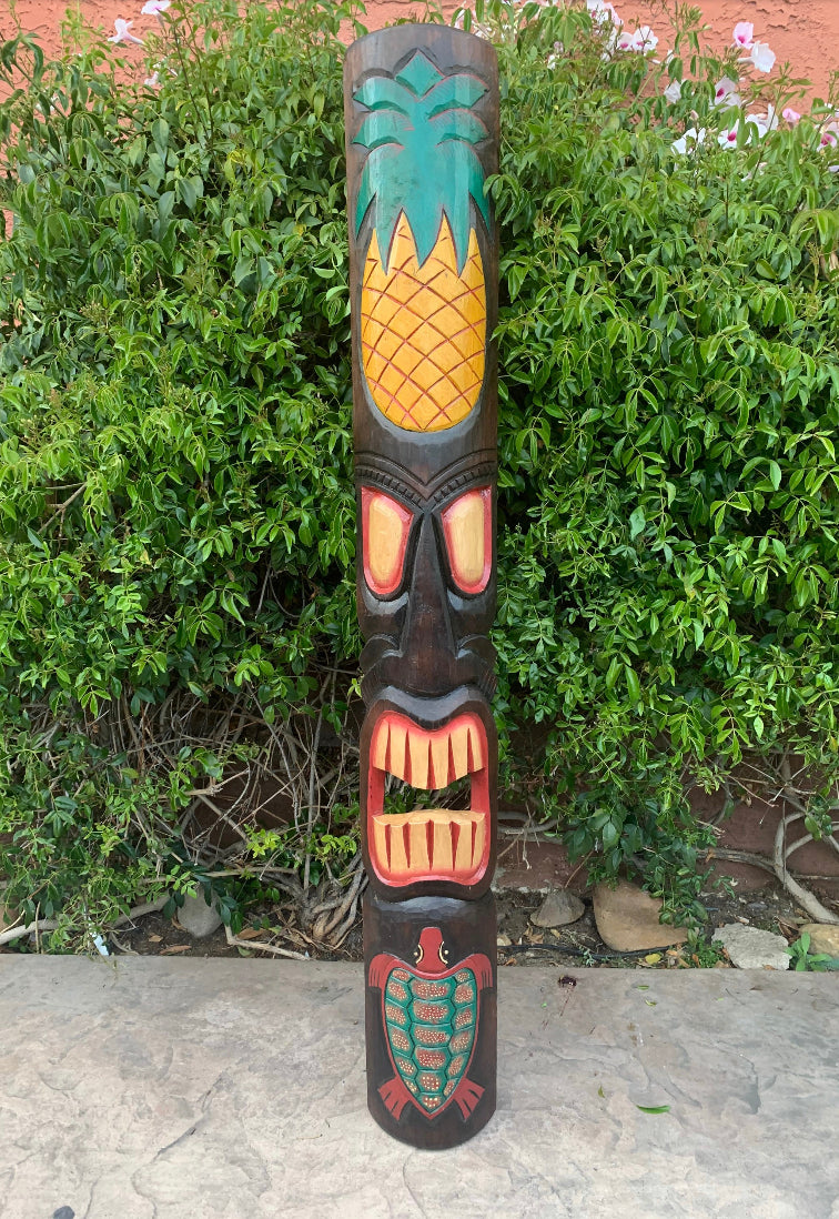 Carved wooden tiki totem with pineapple motif and red mouth in a garden.