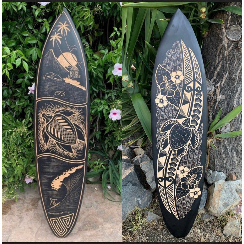 Two black surfboards with tan Polynesian carvings; left shows island and palm trees, right features floral geometric patterns.