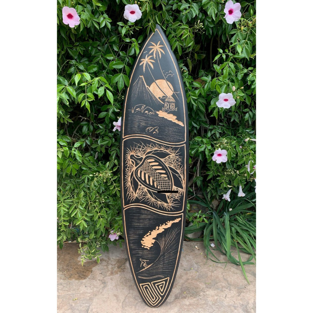 Polynesian carved surfboard with palm trees and waves, resting against a green hedge with pink hibiscus flowers
