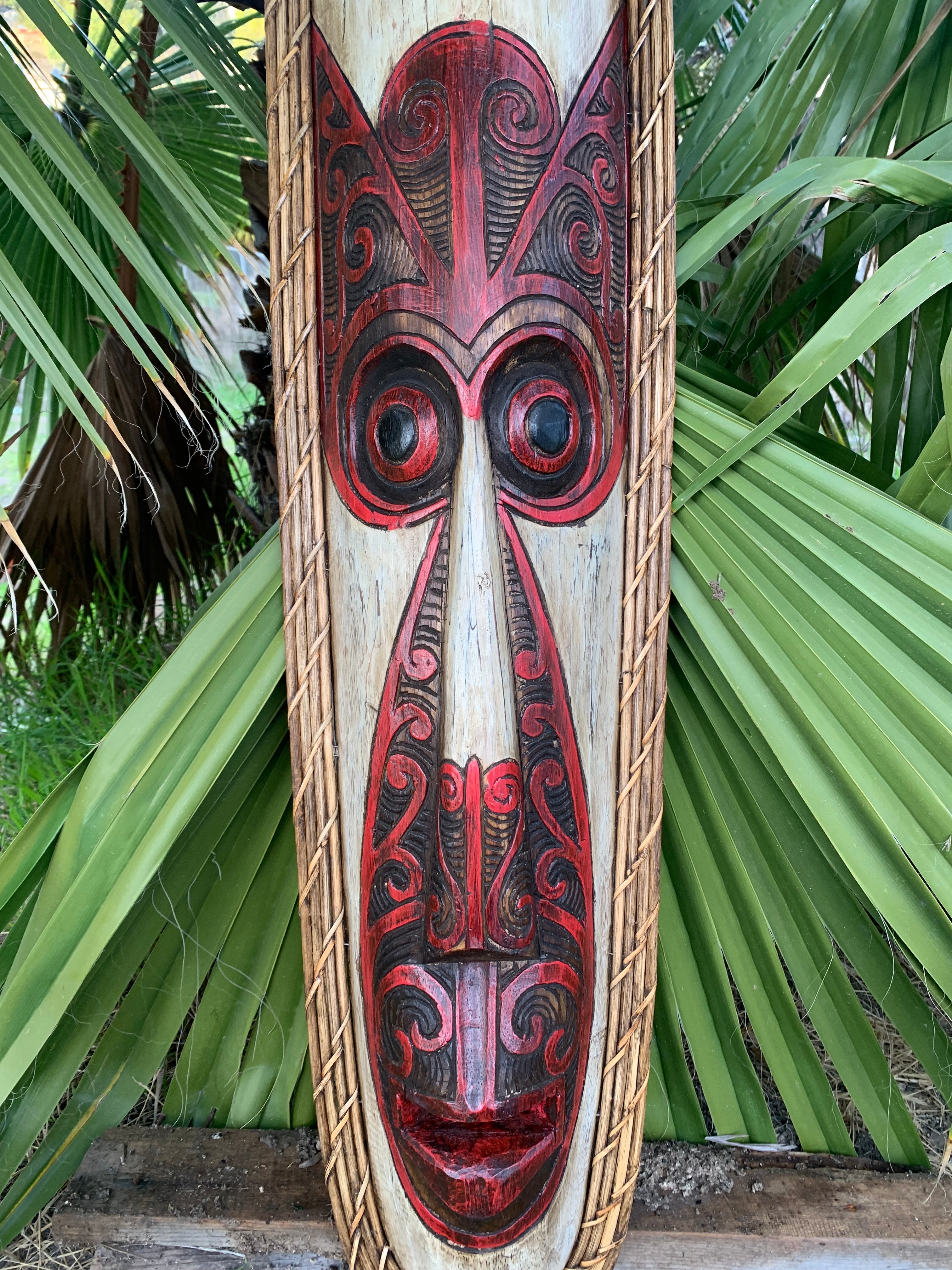 Carved Polynesian tiki mask on a wooden plank with red and black swirl patterns, rope border, palm fronds in the background.