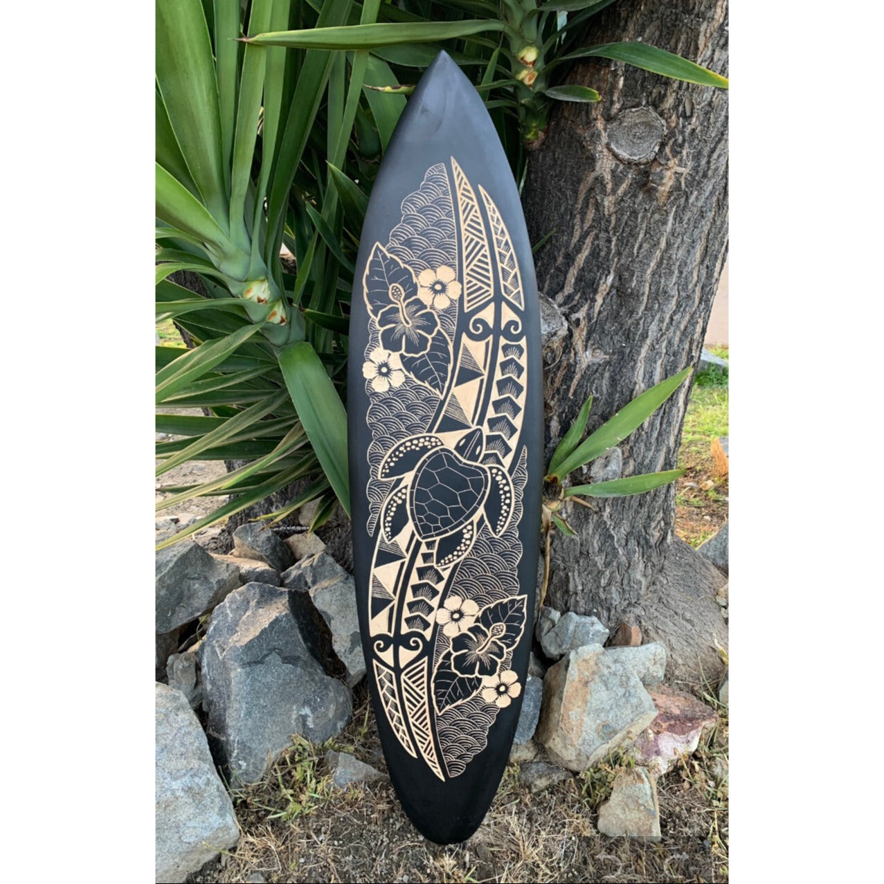 Polynesian tiki-inspired surfboard with floral patterns leaning against a tree.