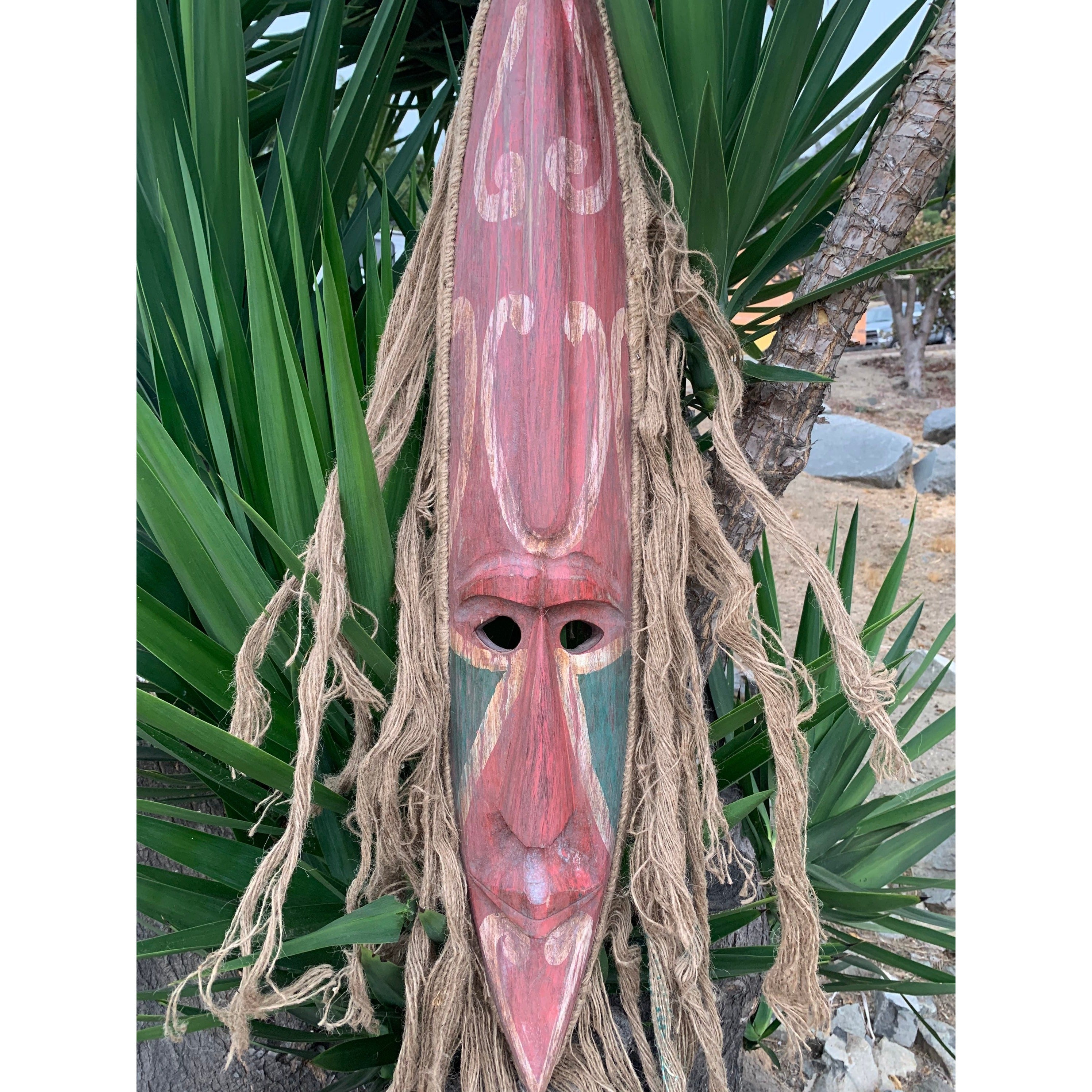 Elongated red wooden tiki mask with rope fringe hanging outdoors among green plants