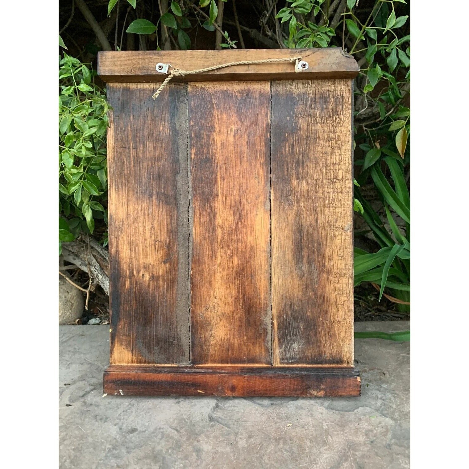 Weathered wooden cabinet with a rope handle outdoors, greenery in the background