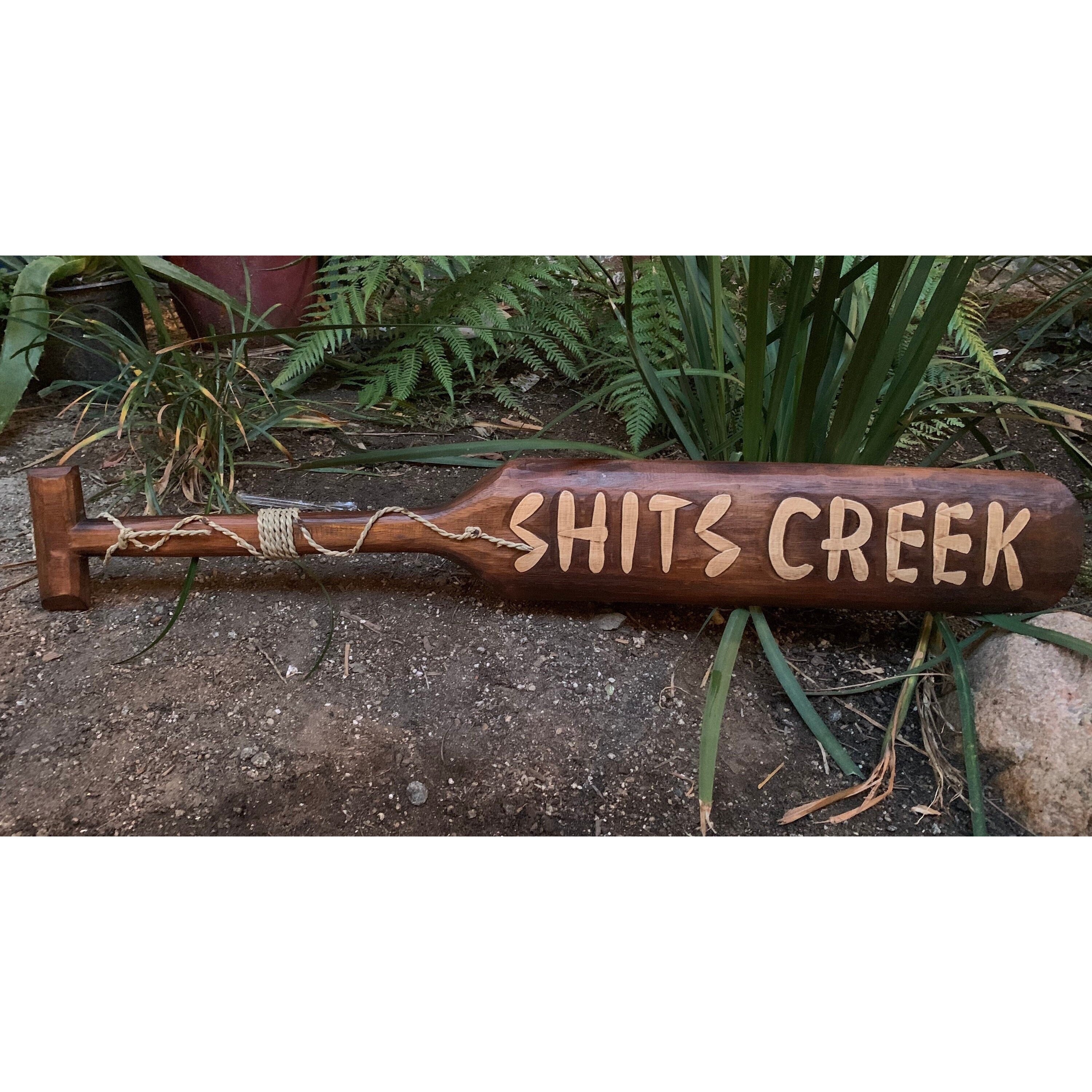 Carved wooden sign reading Shit's Creek laid on the ground among garden plants