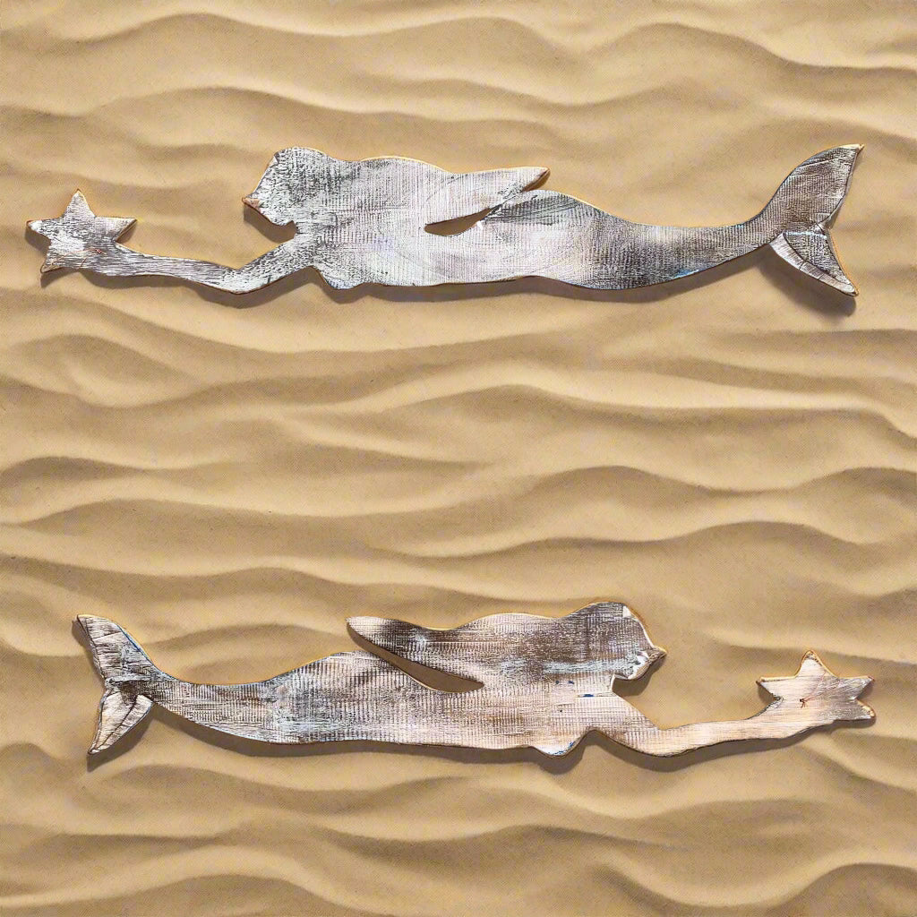 Two silver metal mermaid silhouettes resting on a wavy sandy surface.
