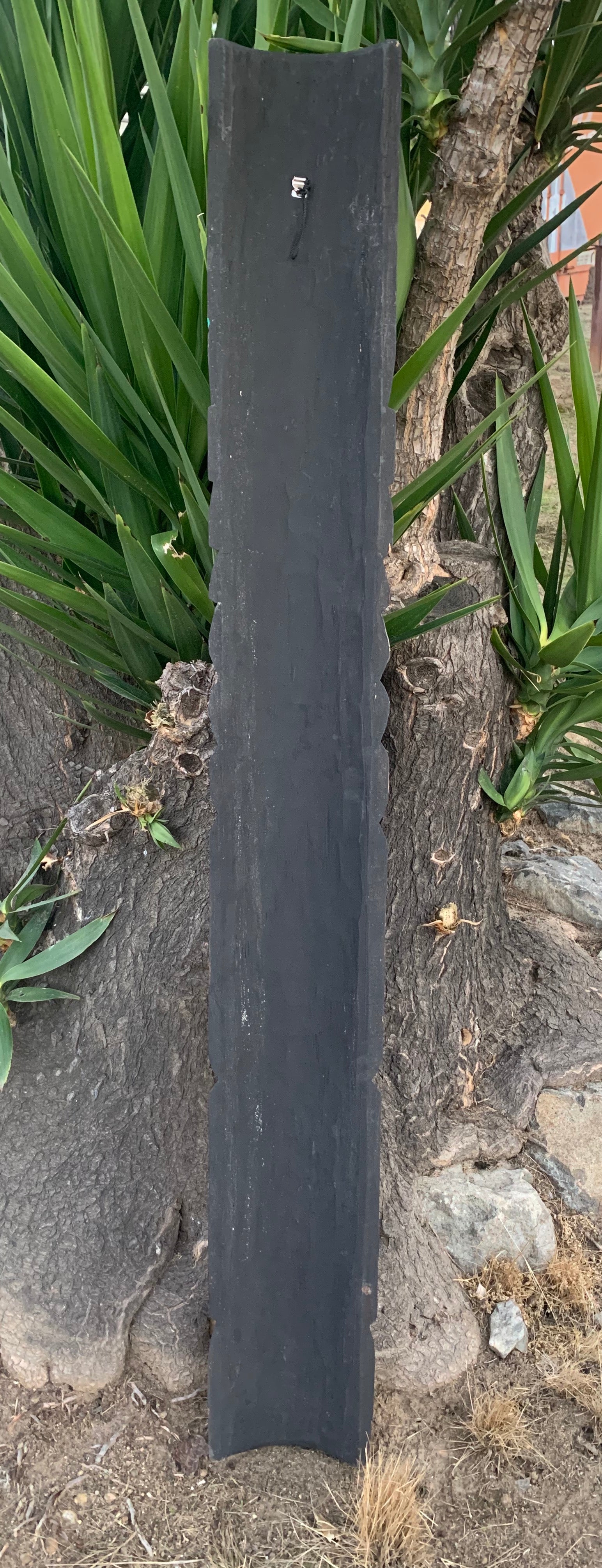 Tall dark slate slab with rough edges leaning upright against a tree in a garden.