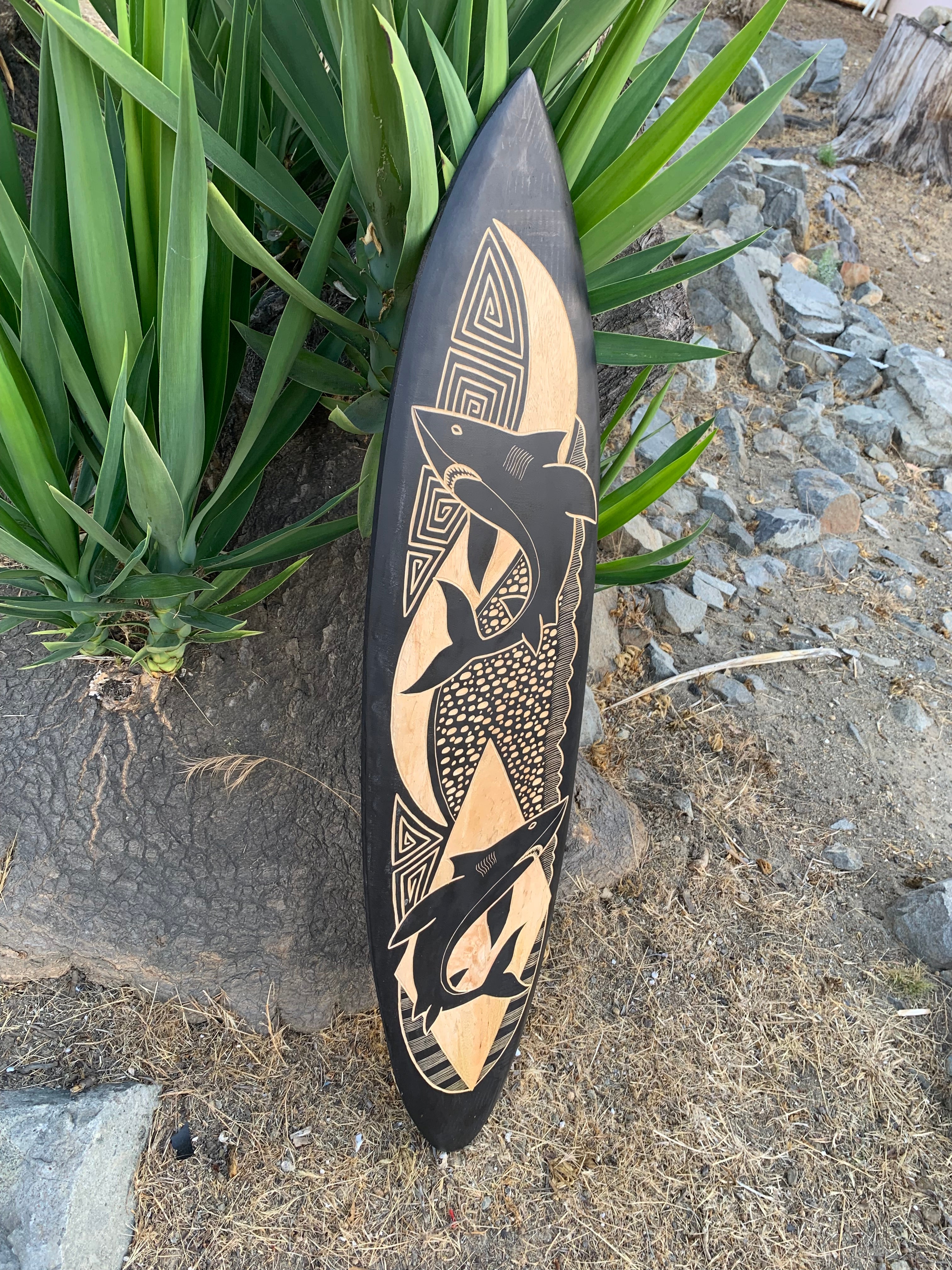 Black surfboard with beige Aboriginal-inspired geometric fish and spiral patterns leaning against greenery