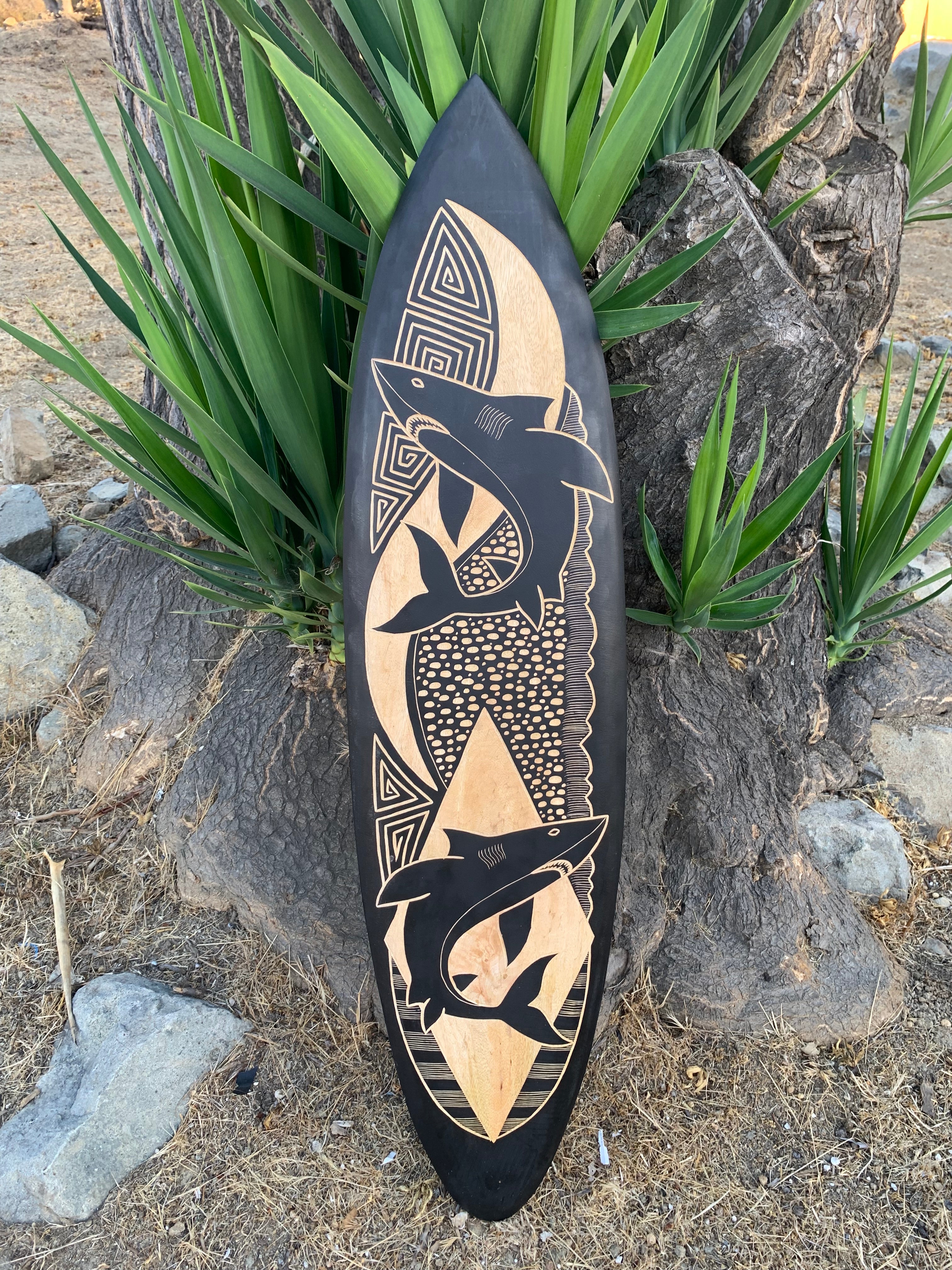 Surfboard with black and natural wood print featuring sharks and geometric shapes leaning against a tree.