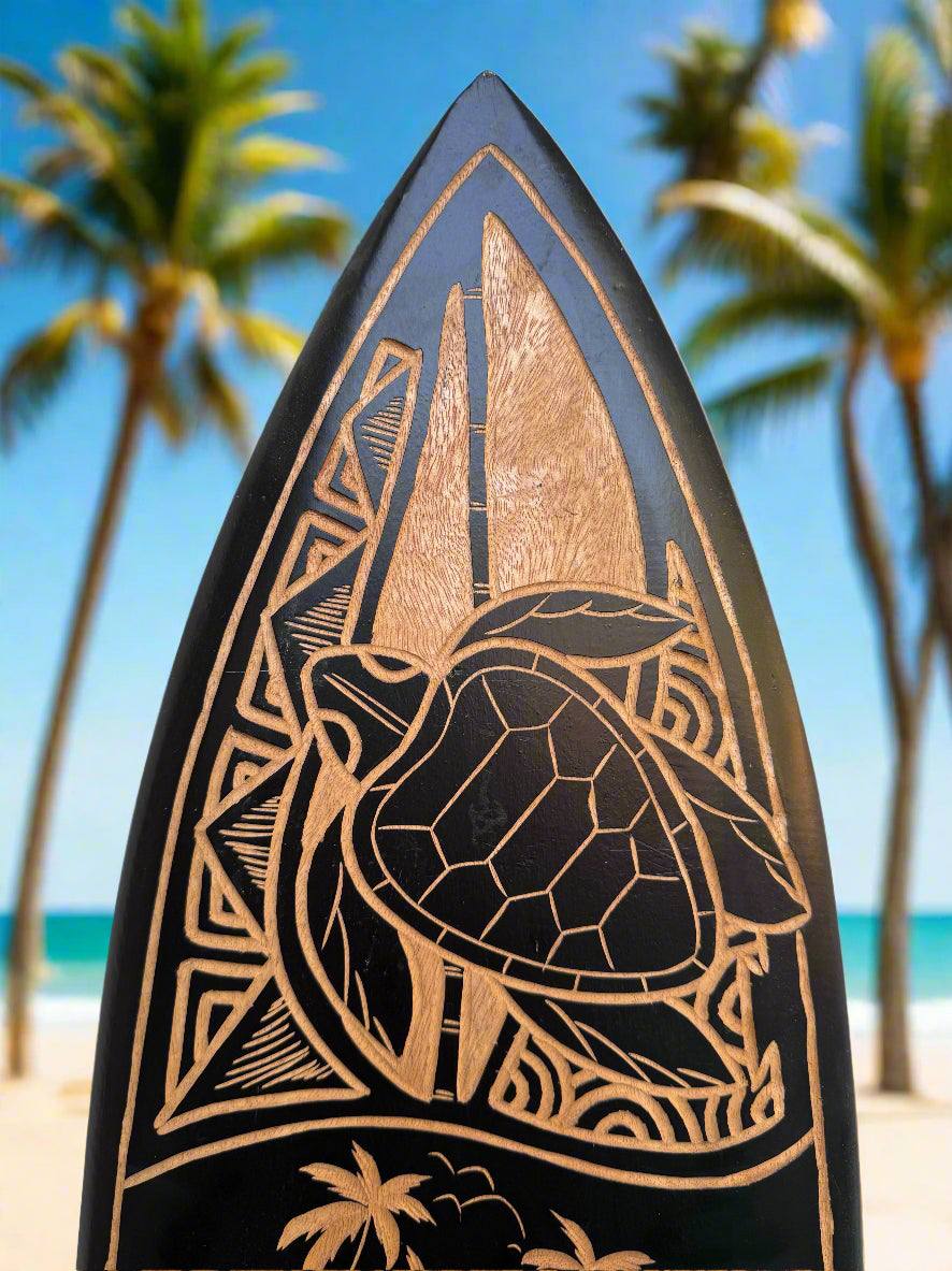 Close-up of a wood-carved turtle design on a surfboard at a sunny beach with palm trees.