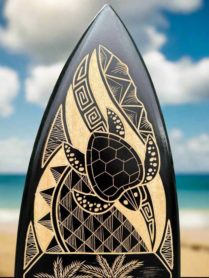Close-up of a black surfboard with beige tribal turtle engraving on the beach under a blue sky.