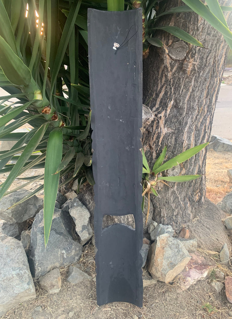 Tall black slate sign with a metal hook, leaning against a tree outdoors; features a curved bottom cutout.