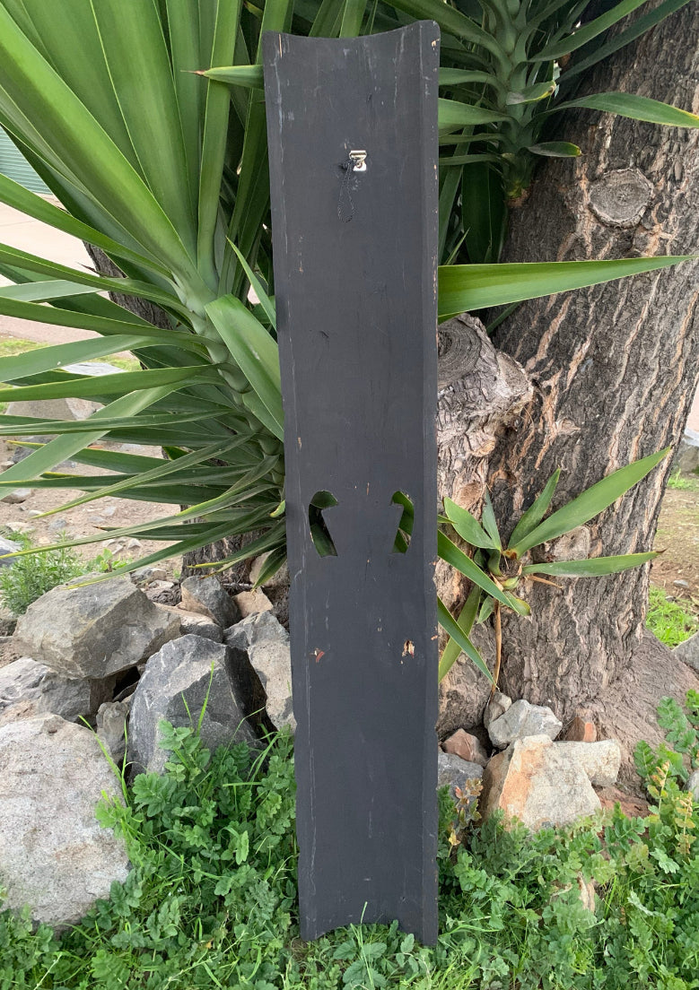 Tall black wooden post with two cutouts leaning against a tree in a garden.