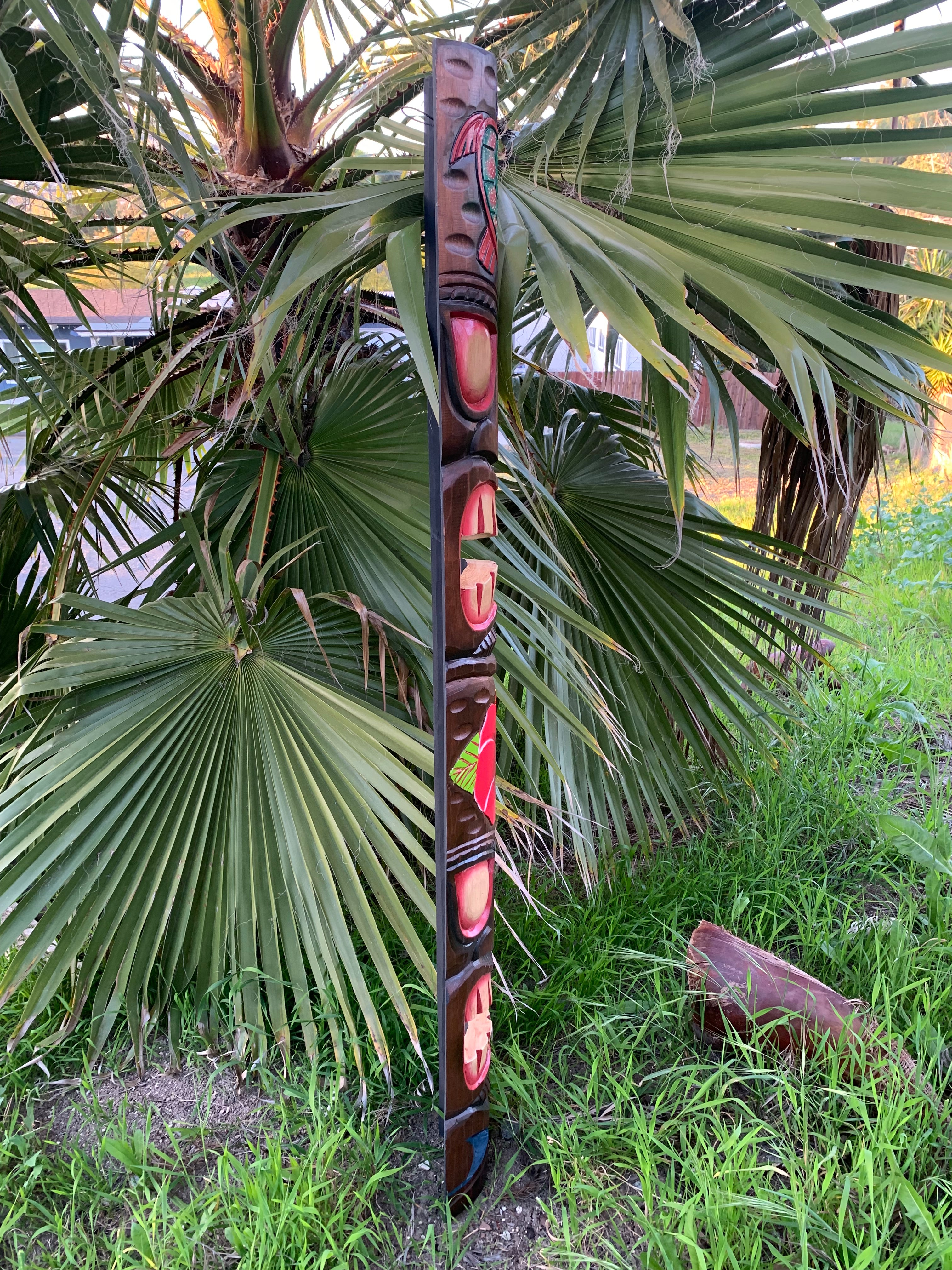 Tall carved totem pole with red inlays beside palm leaves in a garden