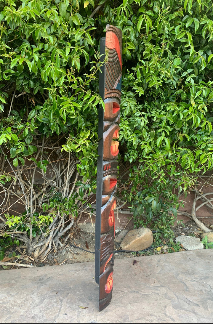 Tall carved wooden totem pole with red and blue face carvings leaning against a green hedge