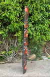 Tall carved wooden totem pole with red and blue face carvings leaning against a green hedge