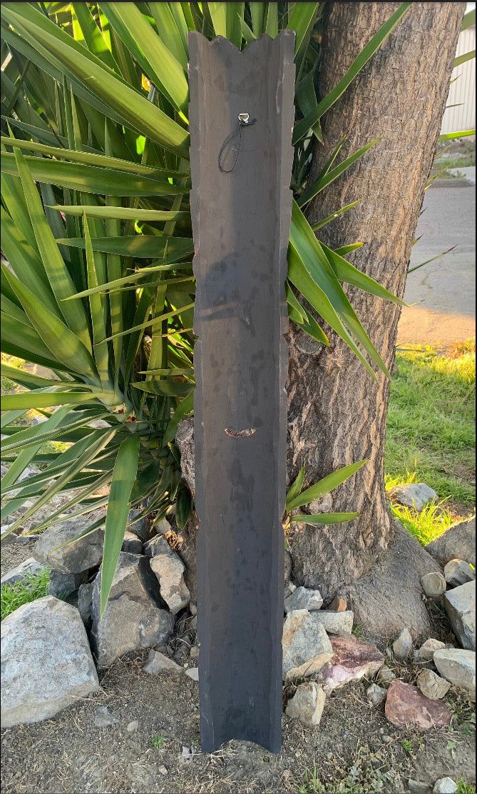 Tall dark metal post with jagged top beside a tree and lush green plants.