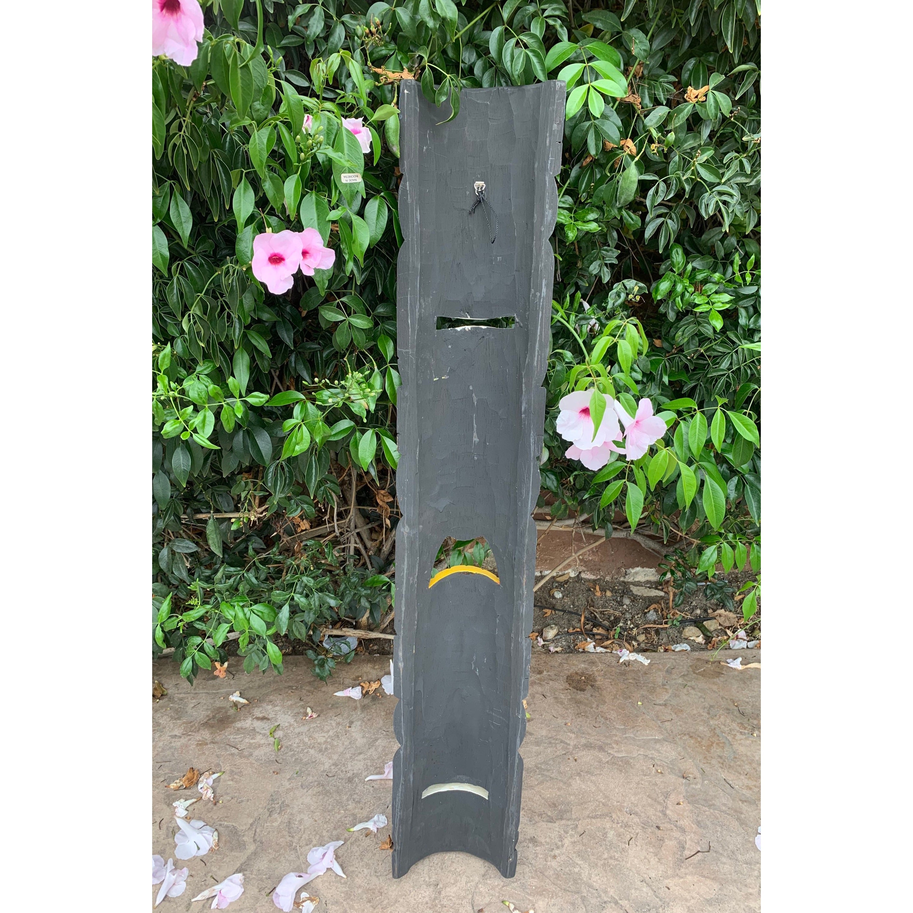 Tall dark wooden sculpture with slotted cutouts in a garden, surrounded by pink hibiscus flowers and green leaves.