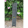 Tall dark wooden sculpture with slotted cutouts in a garden, surrounded by pink hibiscus flowers and green leaves.