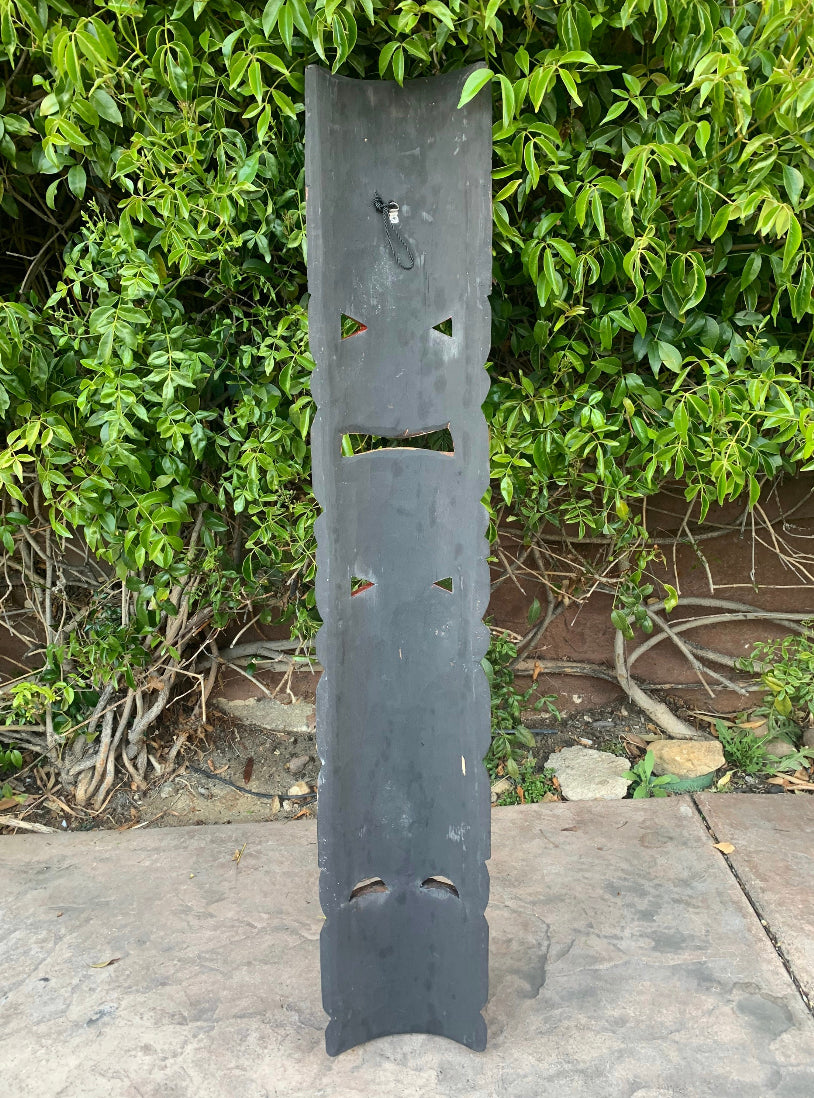 Tall dark metal totem sculpture with face-like cutouts in front of a green hedge