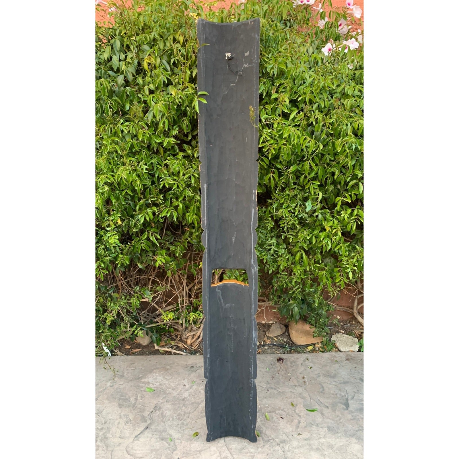 Tall vertical slate slab with a bottom notch, leaning against a green hedge.