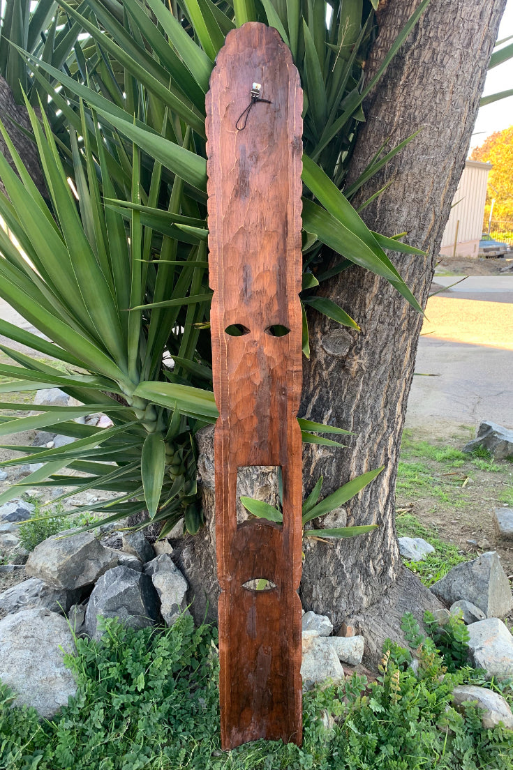 Tall wooden carved mask-like panel with eye holes and rectangular mouth opening leaning against a tree