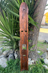 Tall wooden carved mask-like panel with eye holes and rectangular mouth opening leaning against a tree