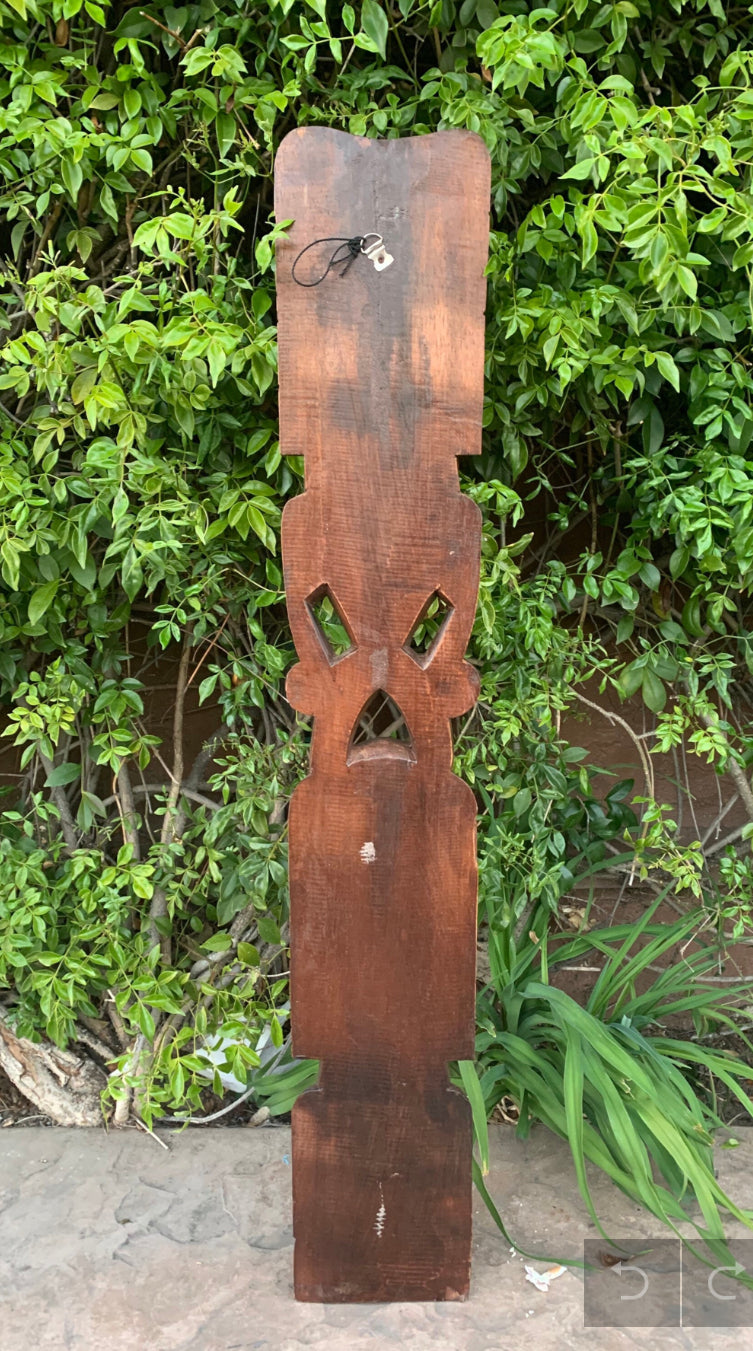 Tall wooden carved totem pole with face cutouts outdoors against green foliage