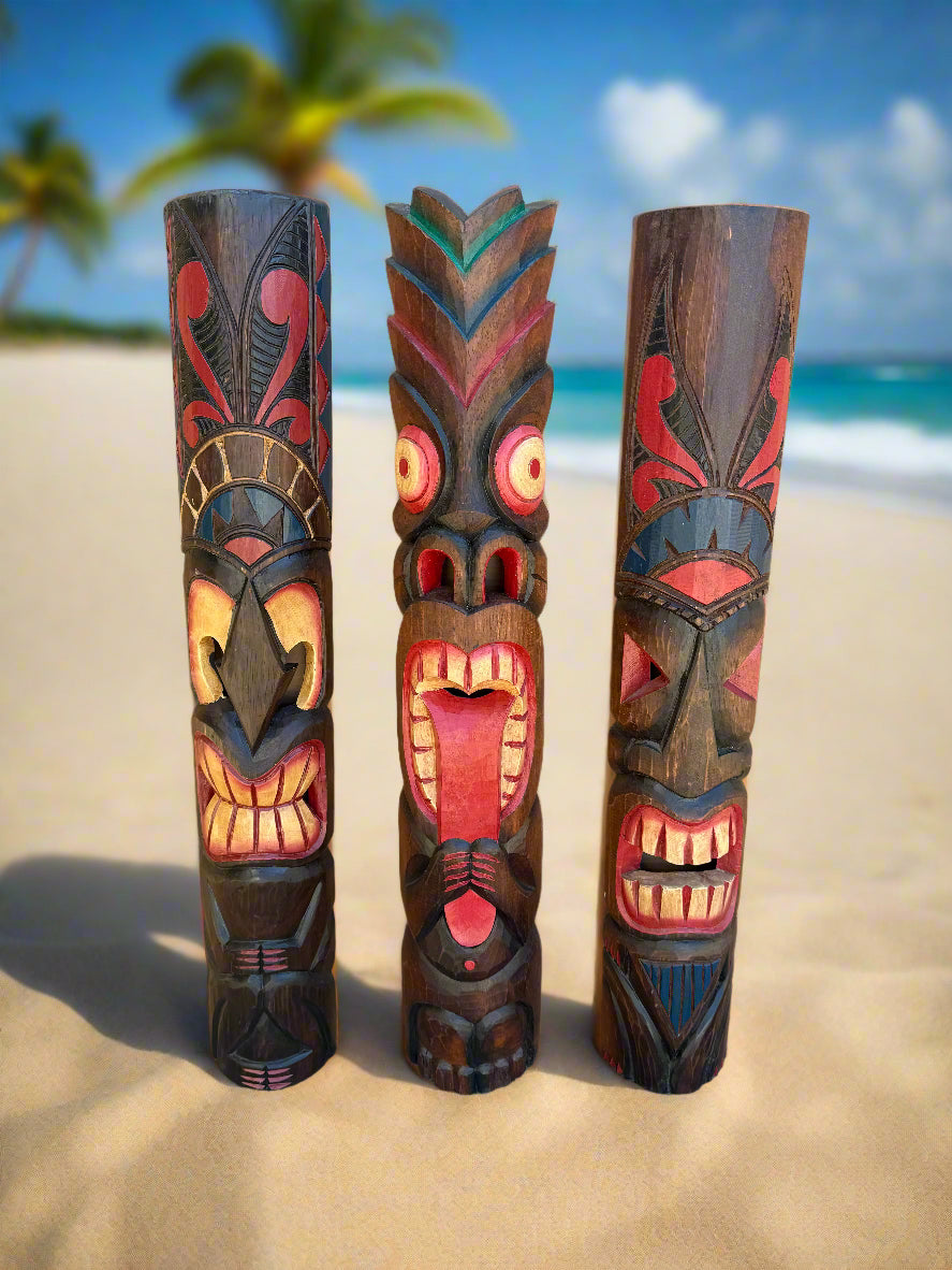 Three carved tiki statues on a sandy beach with palm trees and the ocean in the background.