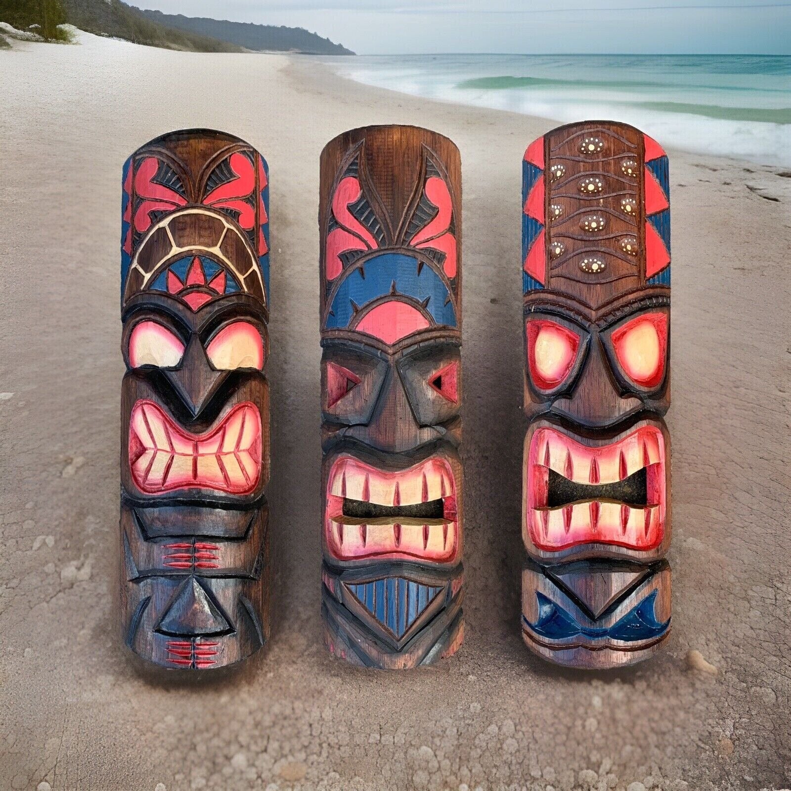 Three colorful wooden tiki totems on a sandy beach with the ocean in the background