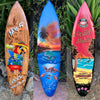 Three decorative surfboards with tropical designs: parrot with cocktail, sunset beach scene, and liquid aloha branding