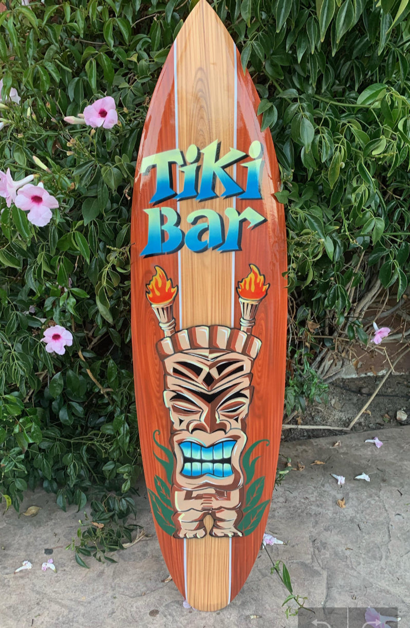 Decorative tiki-bar surfboard with carved tiki face and flaming torches against greenery