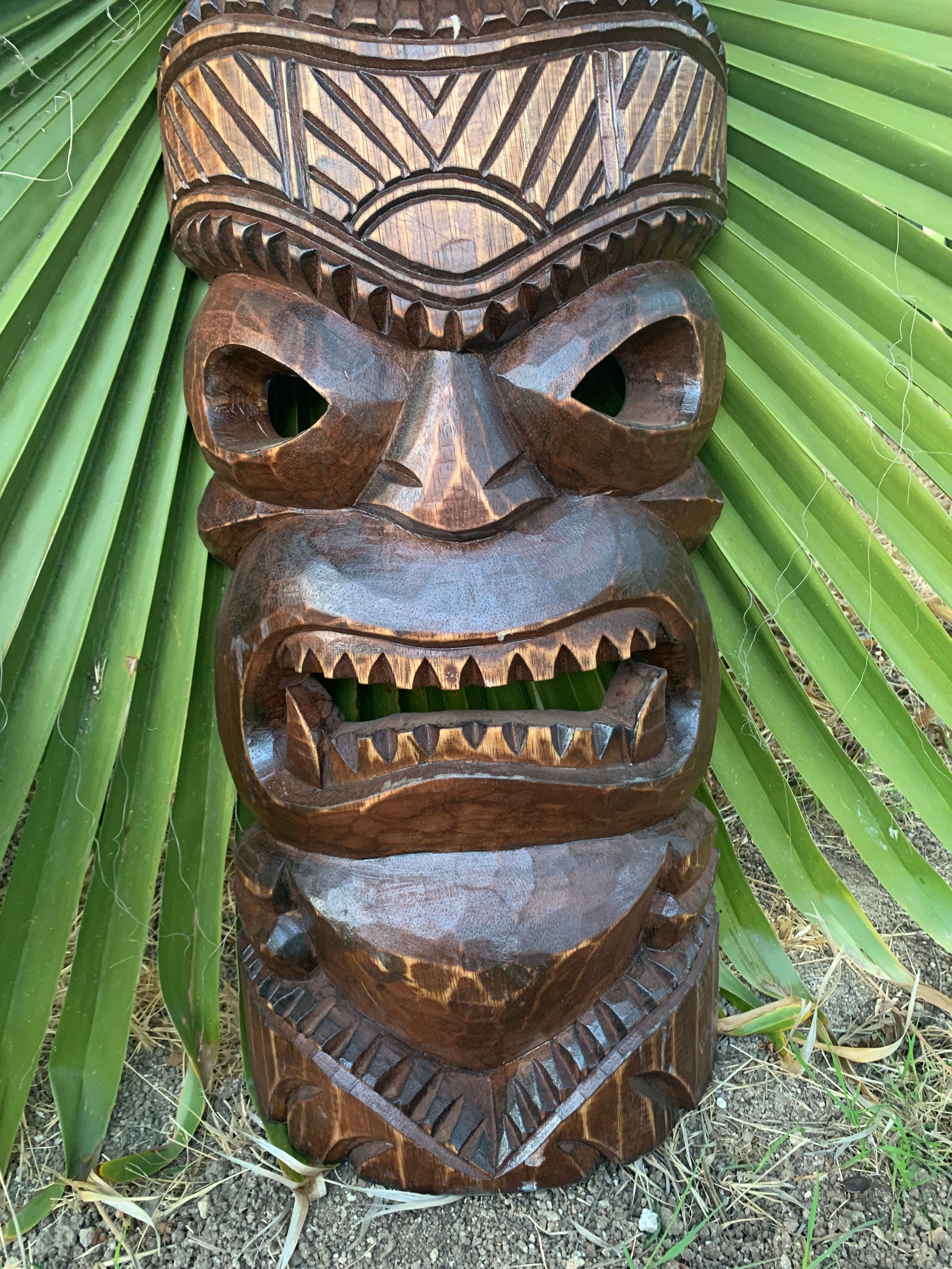 Carved wooden tiki mask with geometric patterns, large hollow eyes and an open toothy mouth against green palm leaves.