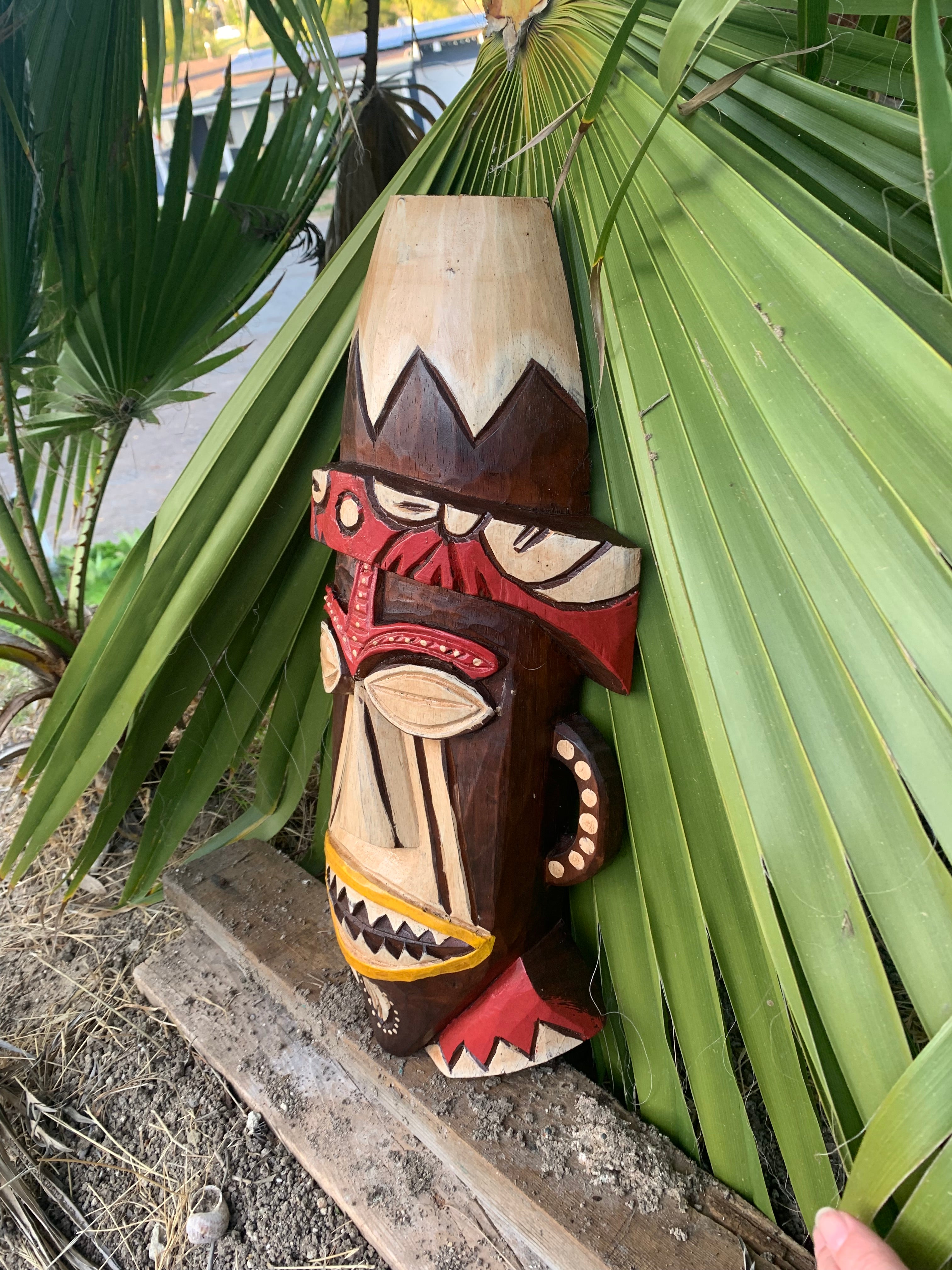 Carved wooden tiki mask leaning against palm leaves outdoors