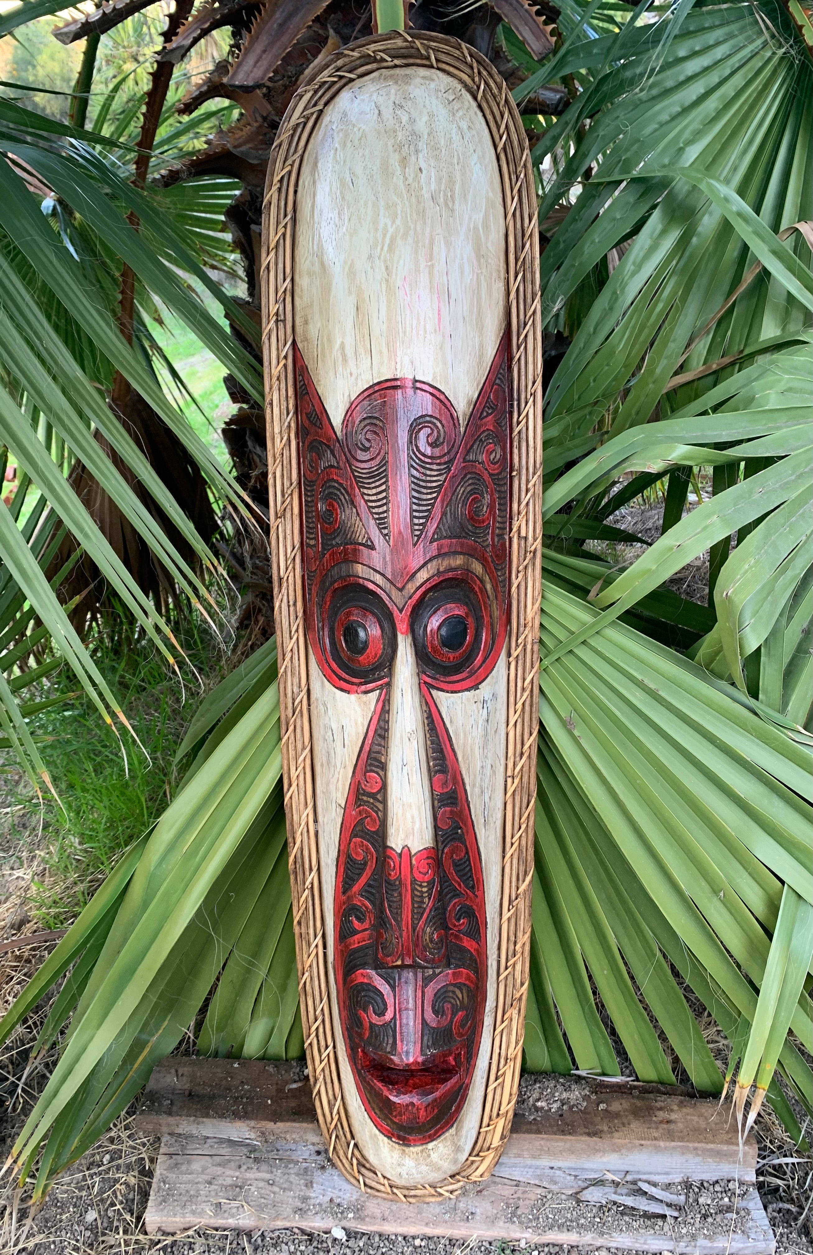 Tall wooden tiki mask panel featuring red and black carved patterns with a rope border outdoors among palm leaves
