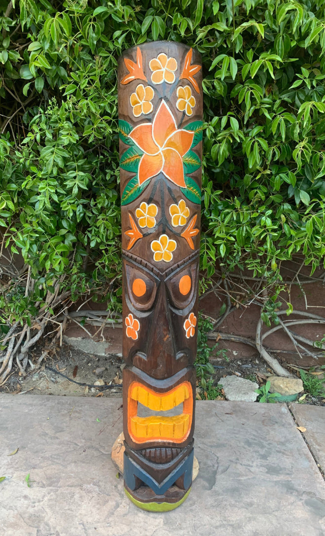 Carved wooden tiki pole with orange flower motifs and a yellow mouth against green foliage.