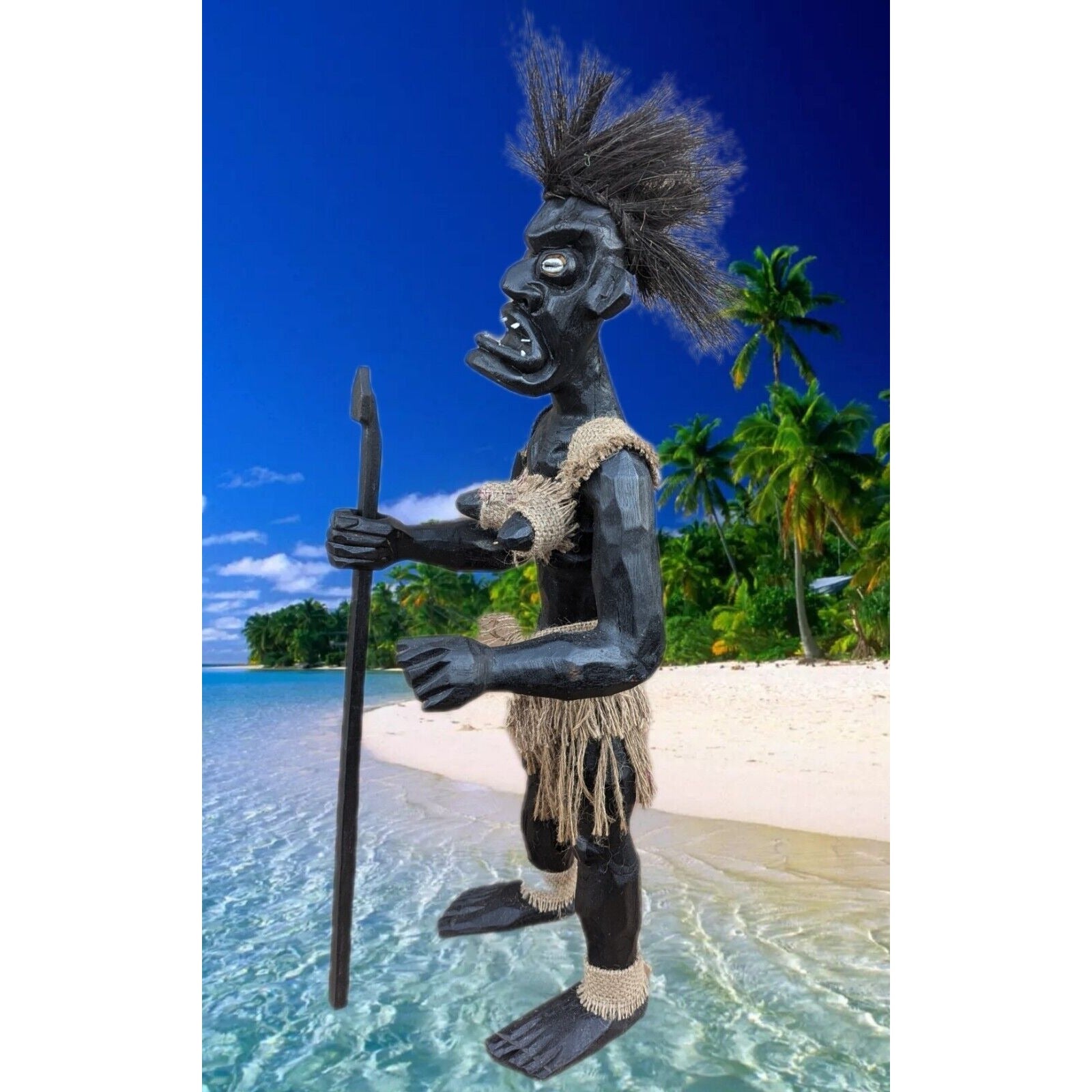Black tiki statue on a tropical beach holding a spear, with palm trees and bright blue sky