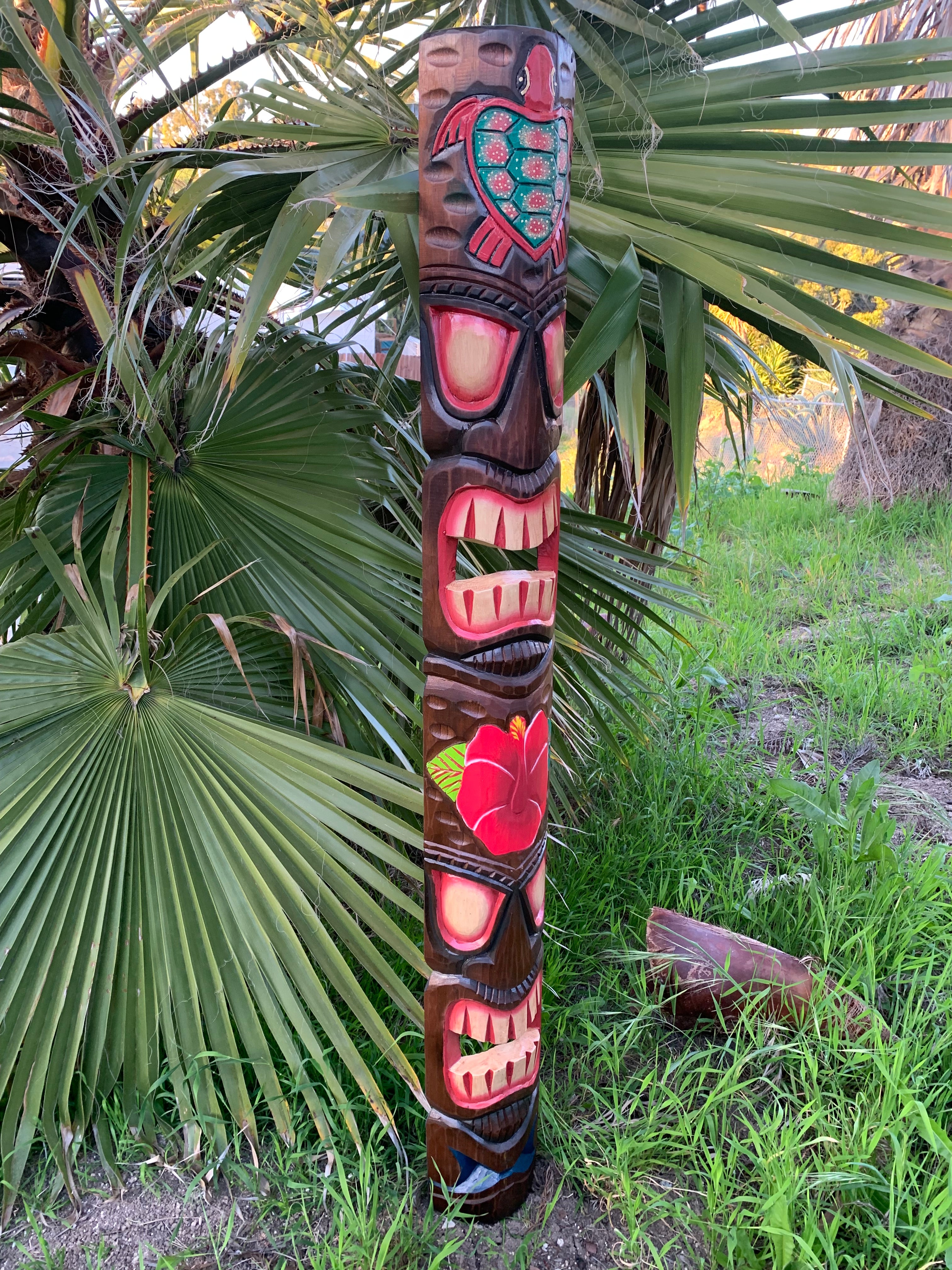 Colorful carved tiki totem pole with face details beside palm leaves in a grassy outdoor setting.