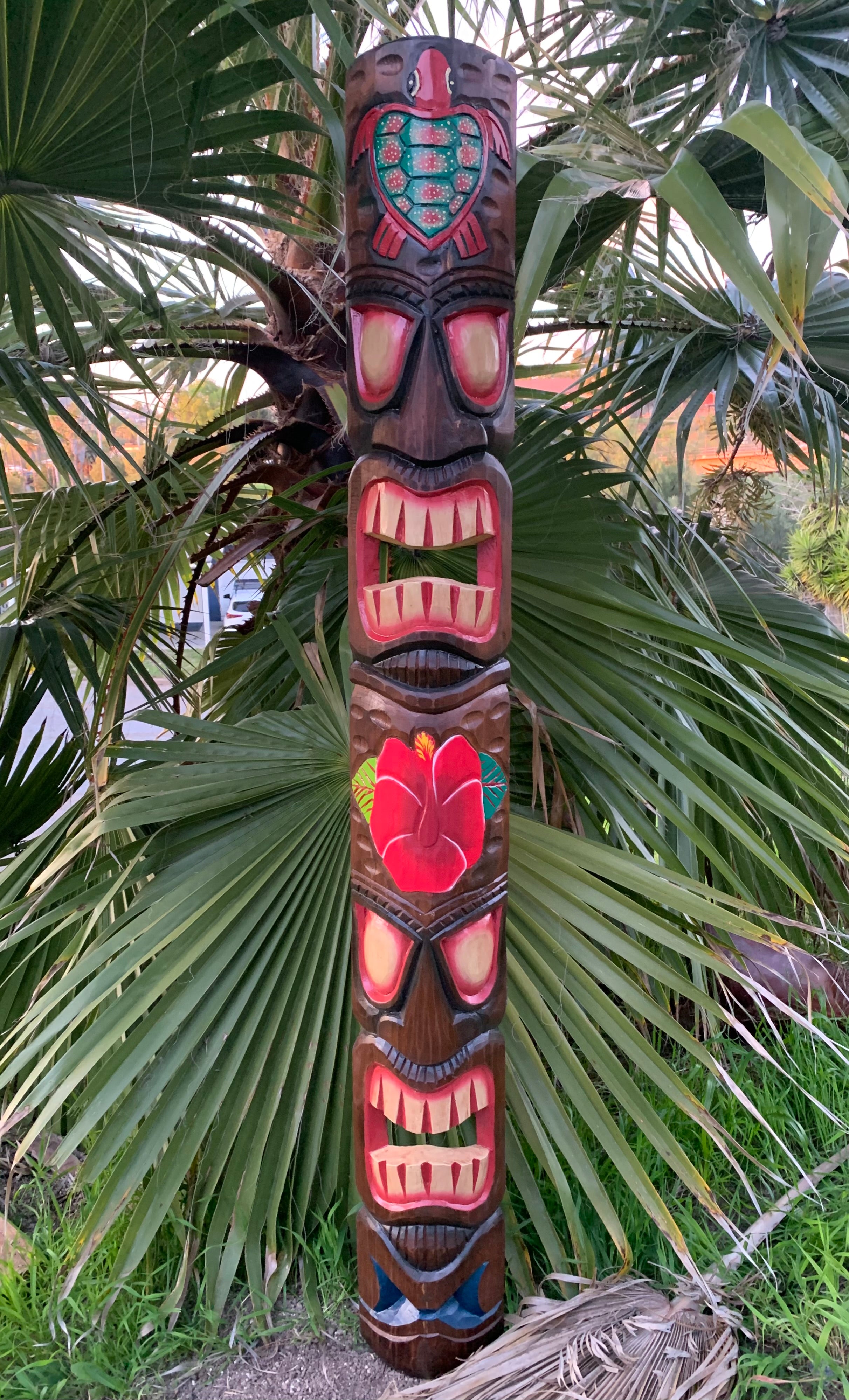 Carved tiki totem pole with colorful faces and hibiscus motif among palm leaves.