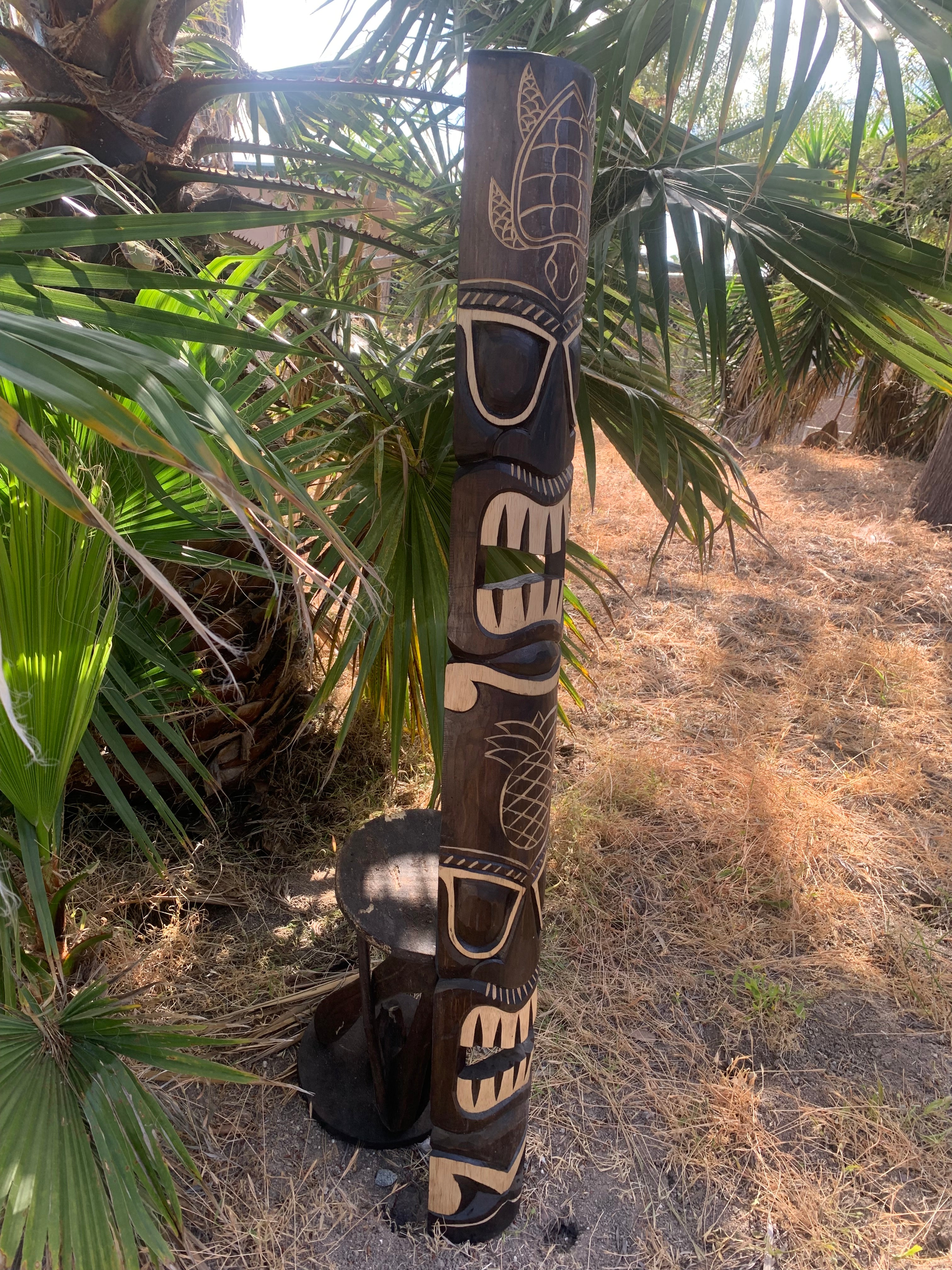 Carved wooden tiki totem pole with facial features in a sunny garden surrounded by palm leaves