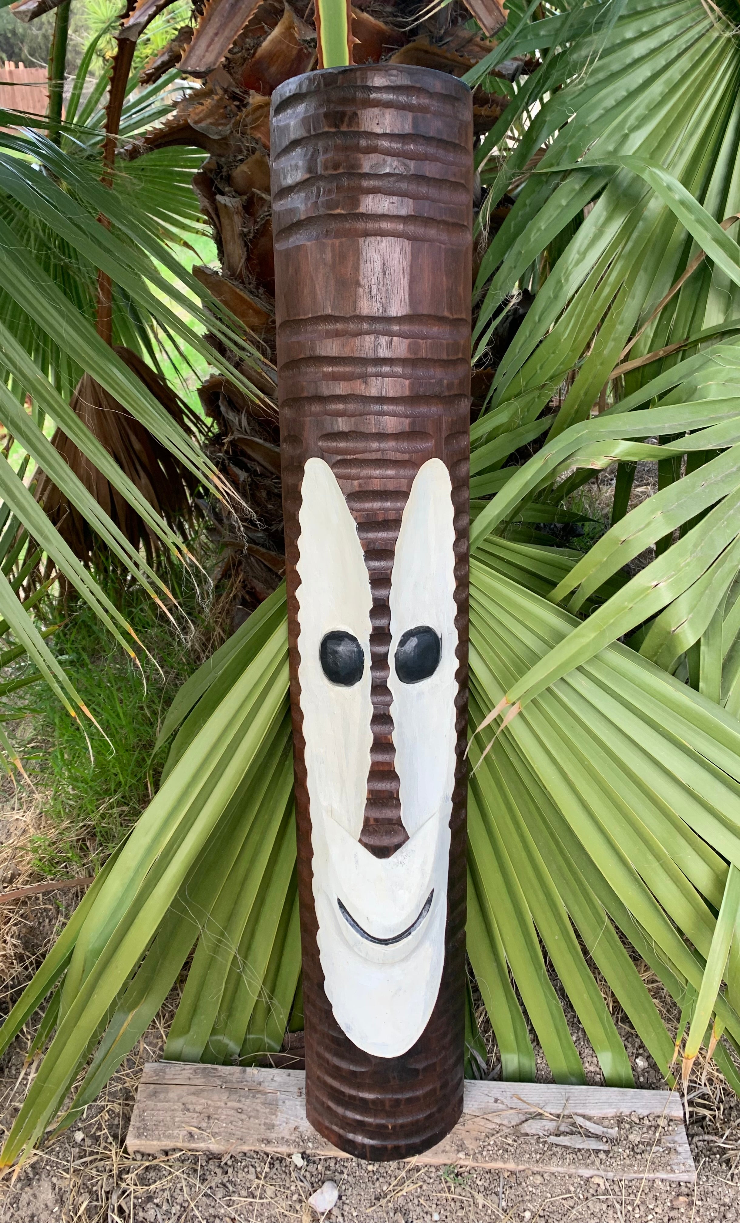 Carved wooden tiki totem pole with a white face and black eyes among palm fronds.