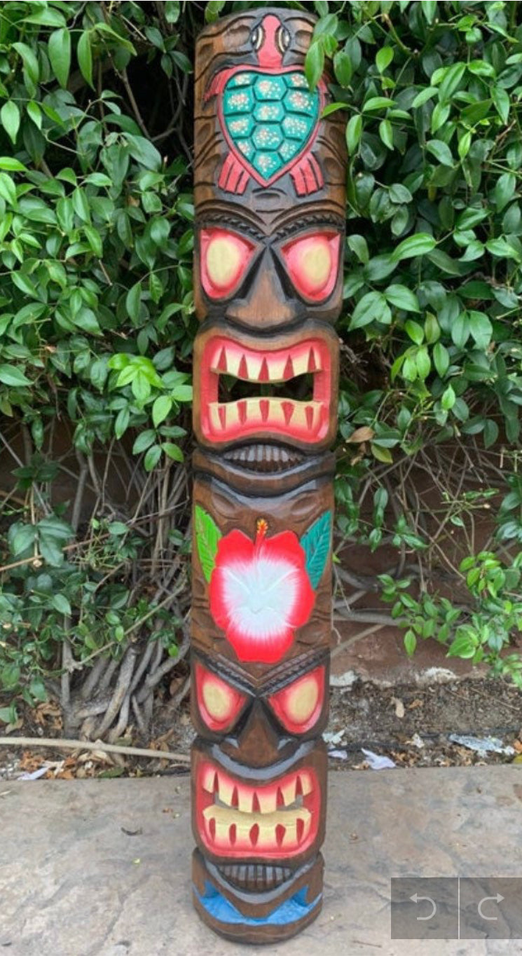 colorful tiki totem pole carving with red facial features and hibiscus flower against lush green foliage