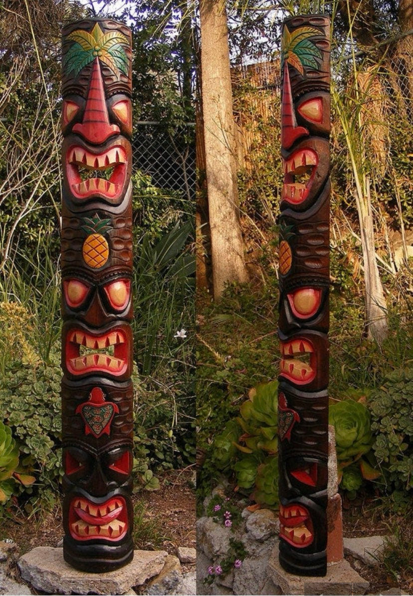 Two carved tiki totem poles with red faces and pineapple motifs in a garden setting.