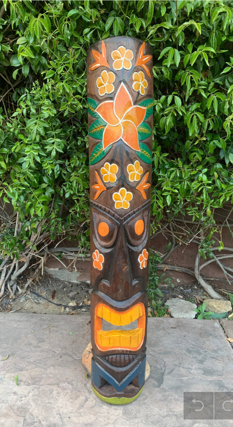 Carved wooden tiki totem with an orange flower, yellow blooms, and geometric facial features amid greenery.