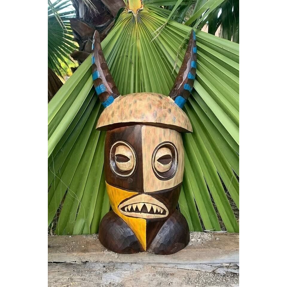 Carved wooden tribal mask with horns, brown and beige panels, geometric eyes, toothy mouth, against palm leaf backdrop