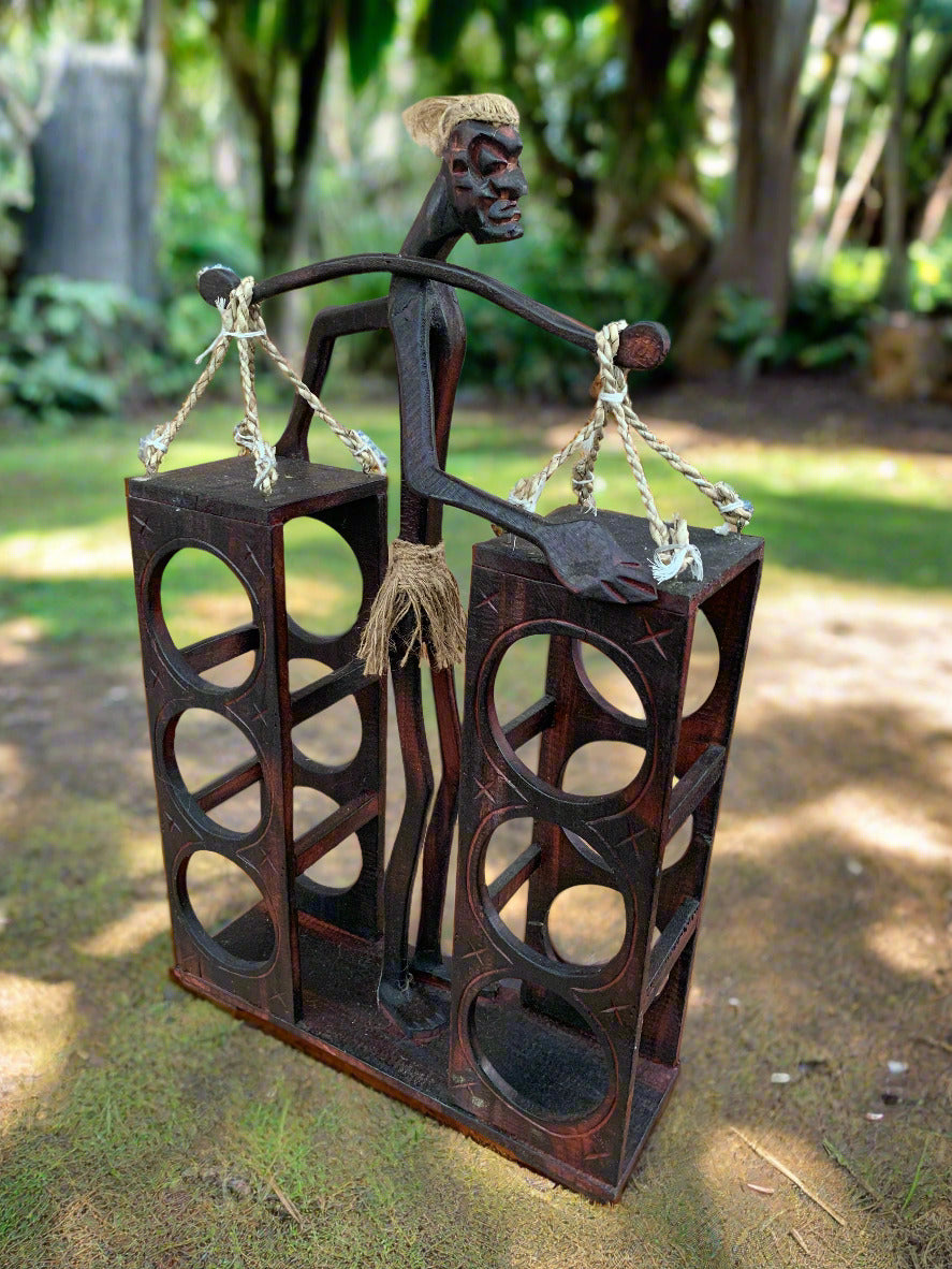 Dark metal tribal figure balancing on two tall speaker-like blocks outdoors